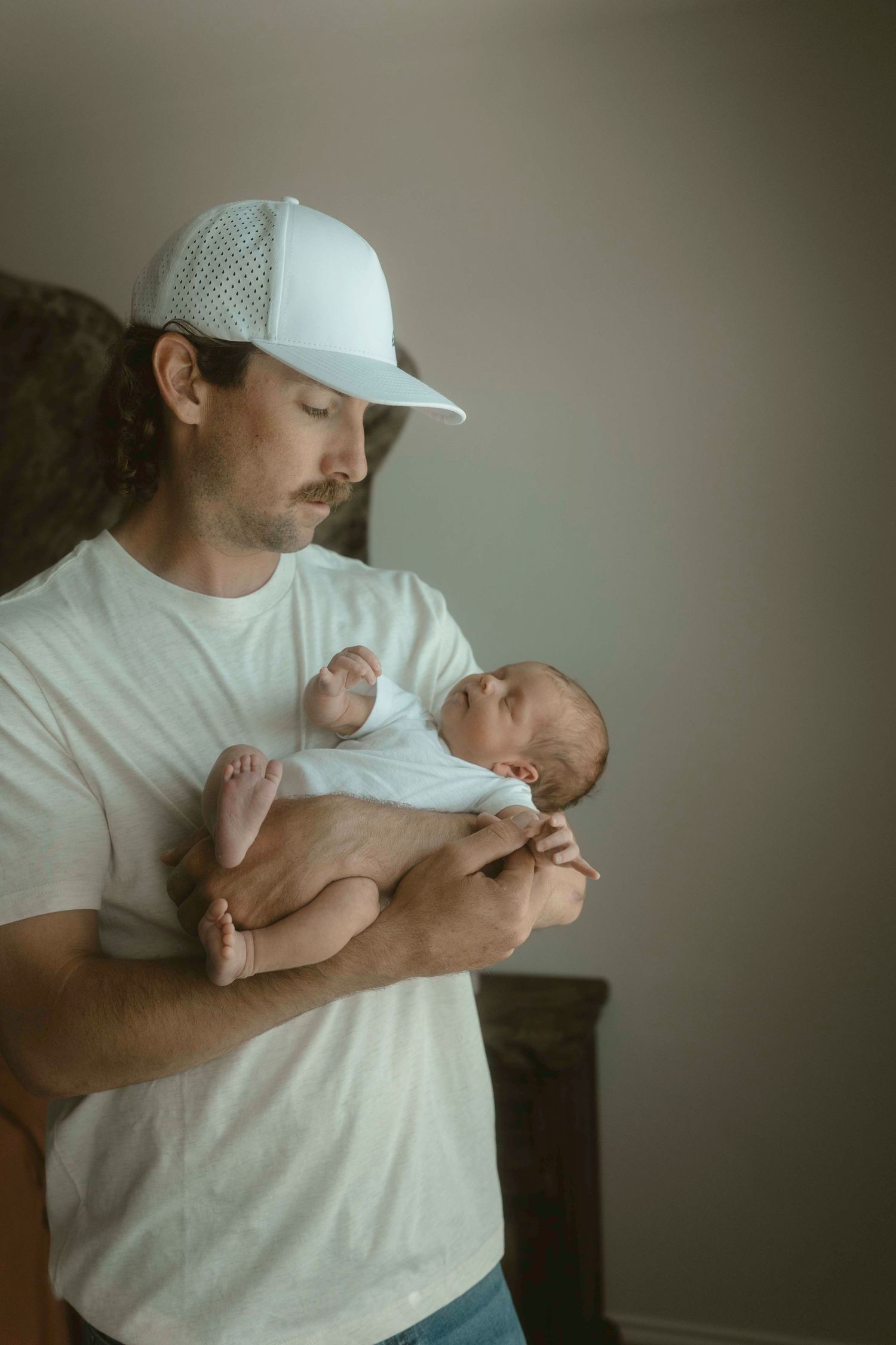 A dad in a white t-shirt and cap tenderly holds and comforts a sleeping newborn baby.