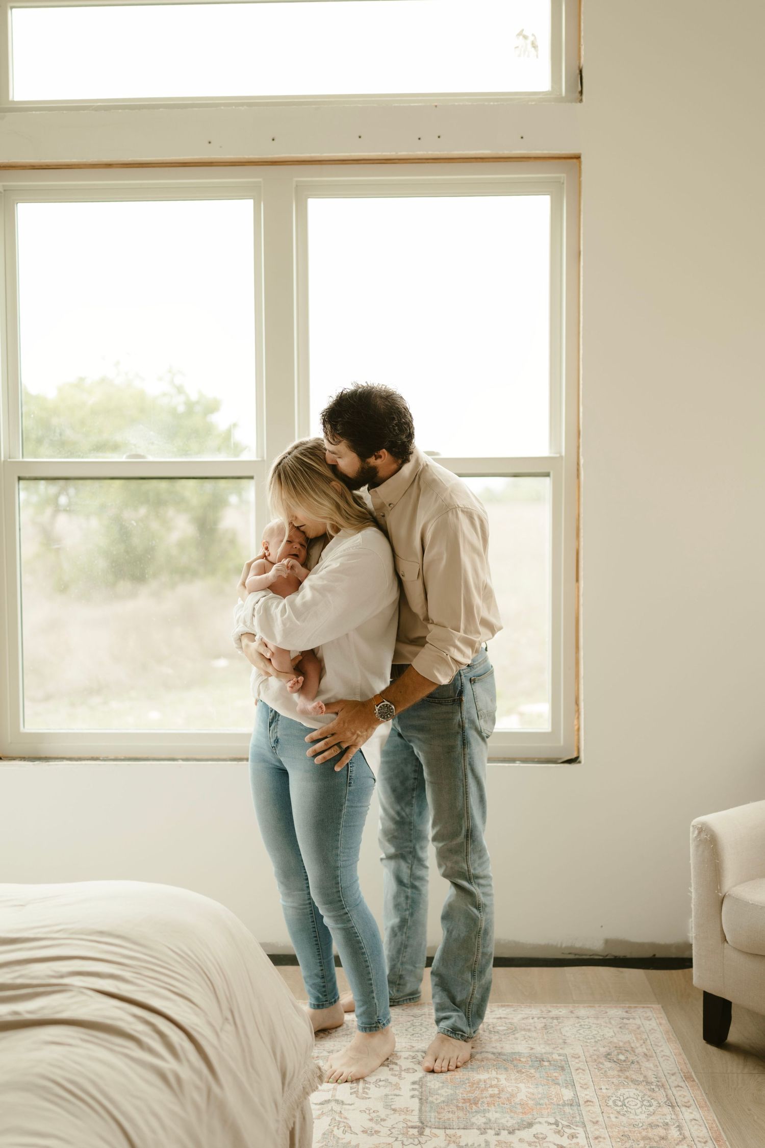 Family shares tender embraces in a bright, sun-lit white room filled with natural light streaming through windows.