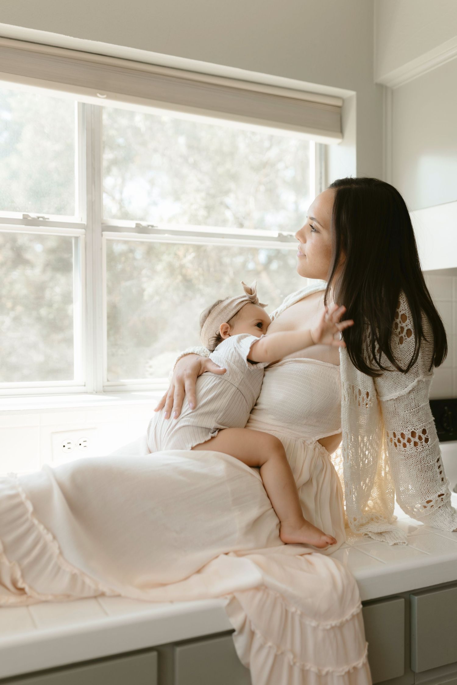 A mother and baby share a tender moment by the window, dressed in white flowing outfits while lounging on a bench seat.