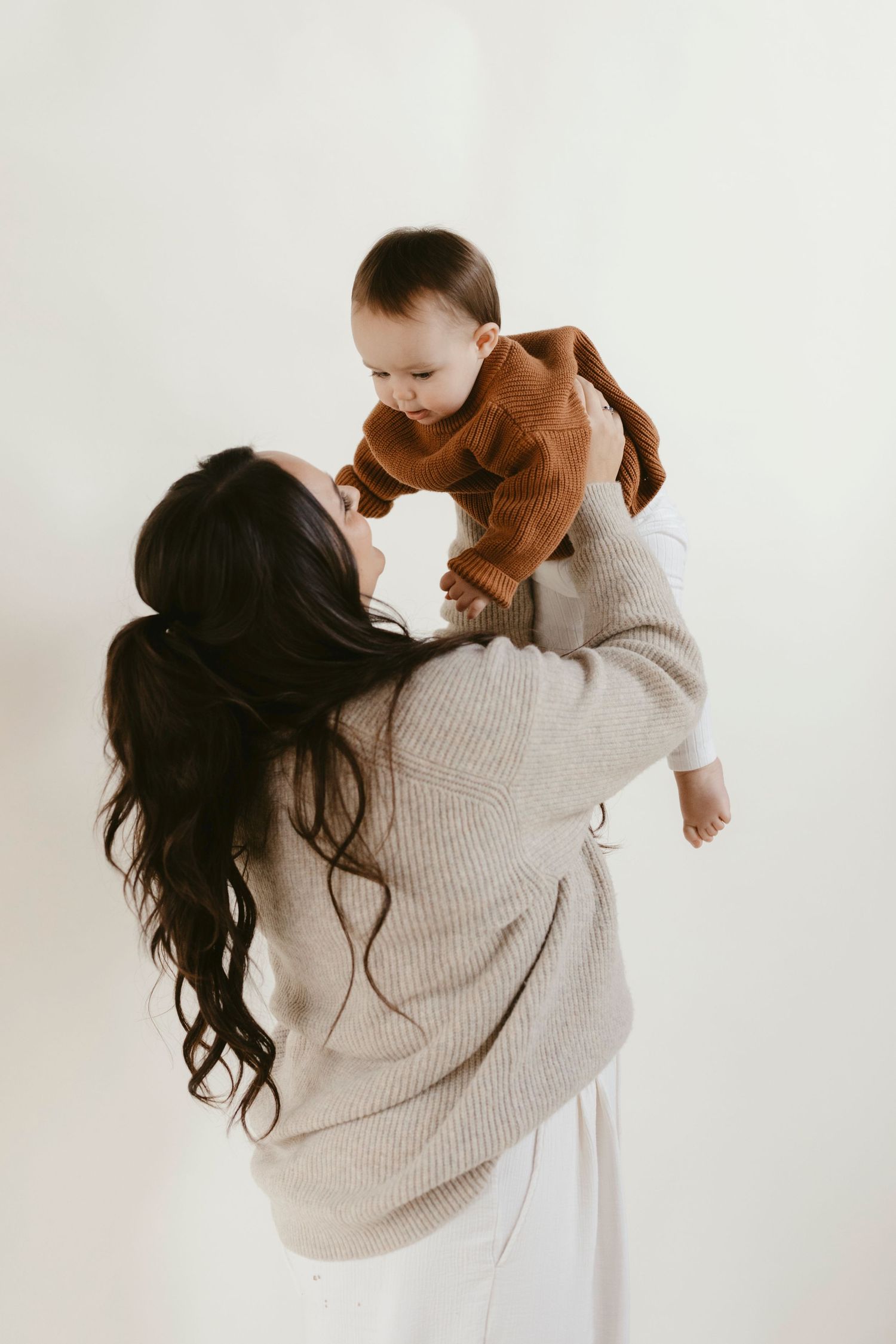 Sweet photo moment between mother and baby in neutral tones as they embrace wearing cozy sweaters against white backdrop.