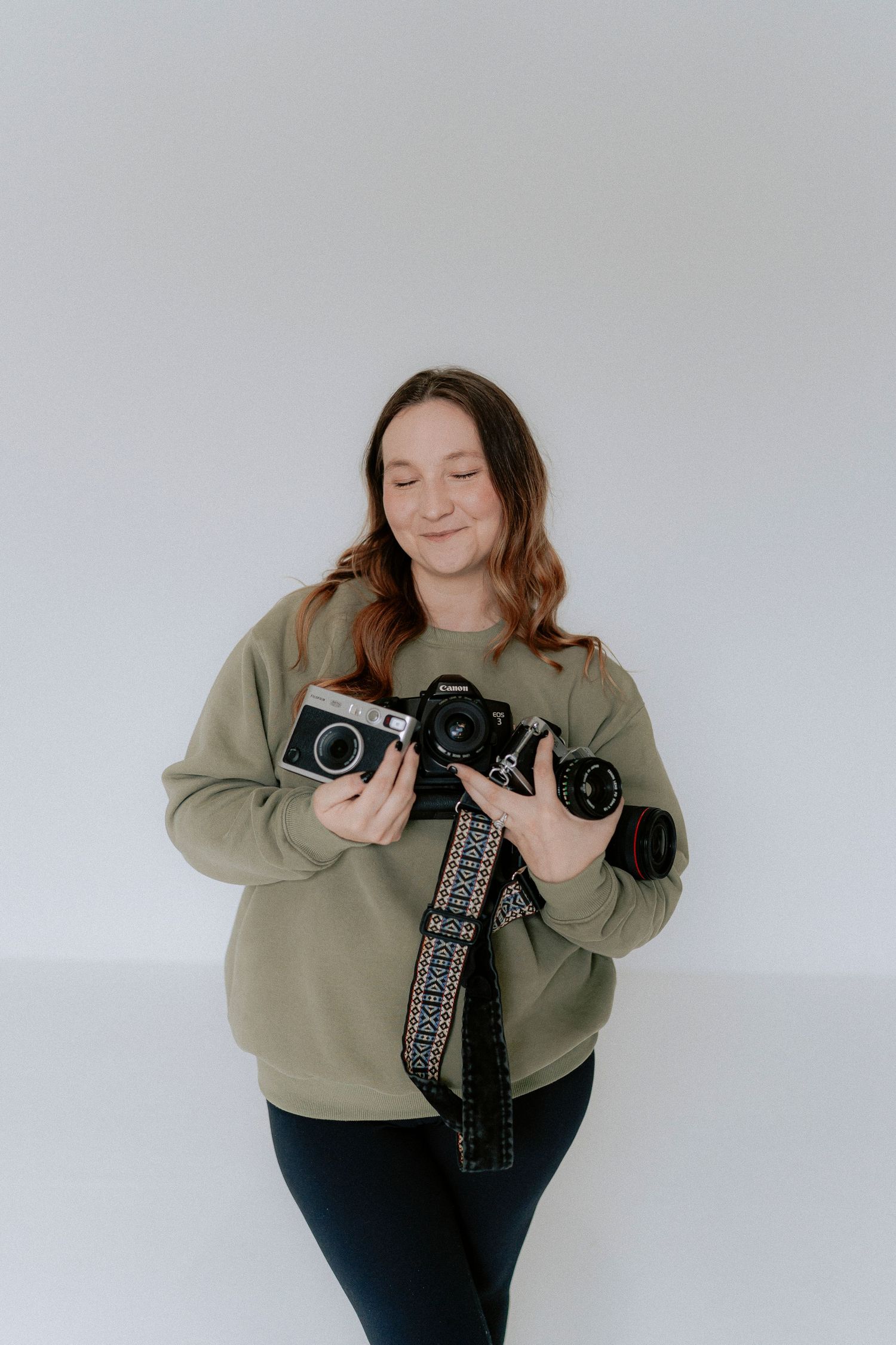 A photographer in a green sweatshirt and dark pants holds vintage cameras while posing against a white background.