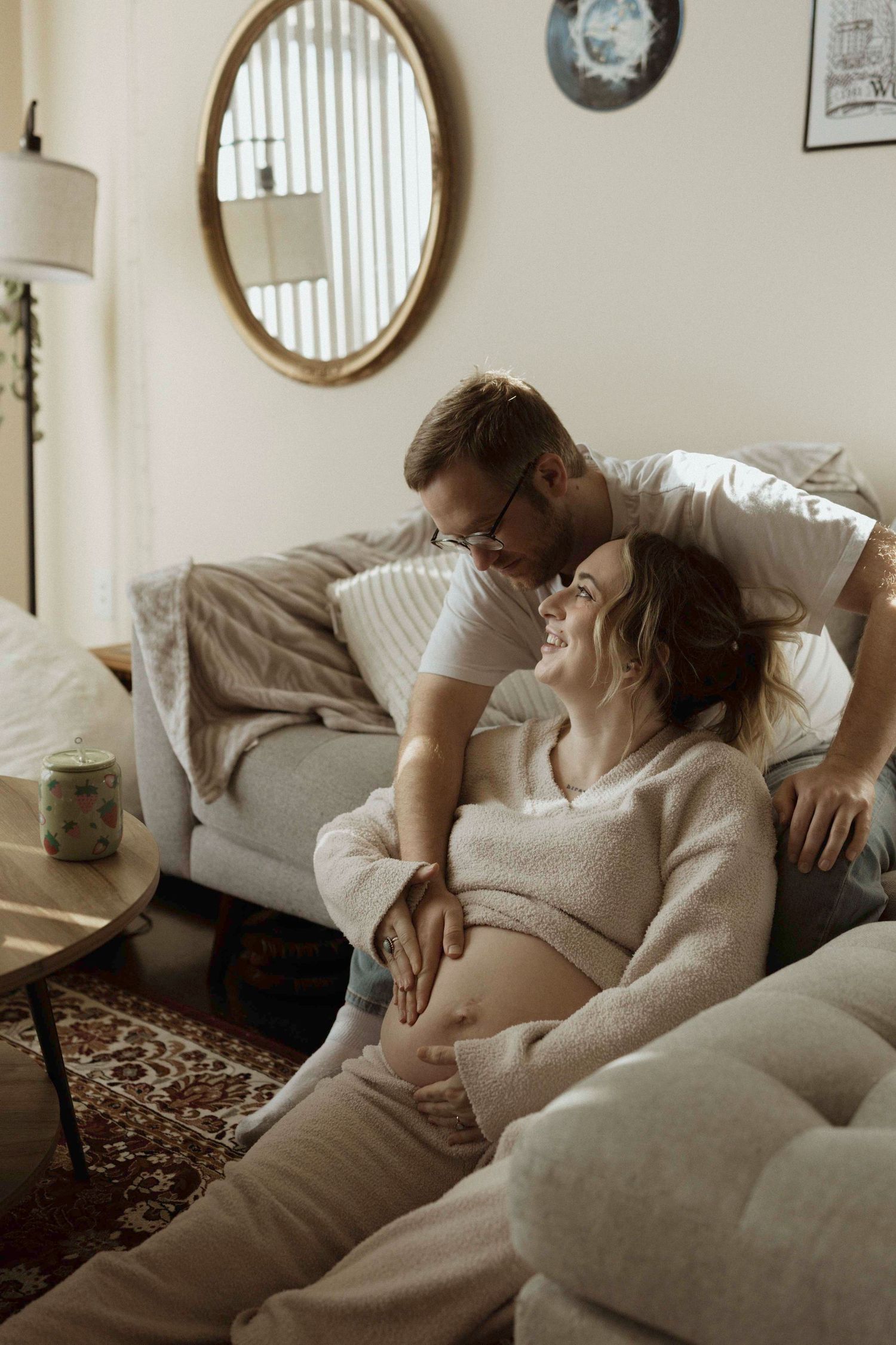 A dad sitting on the couch, pregnant mom sitting on the ground admiring her belly.