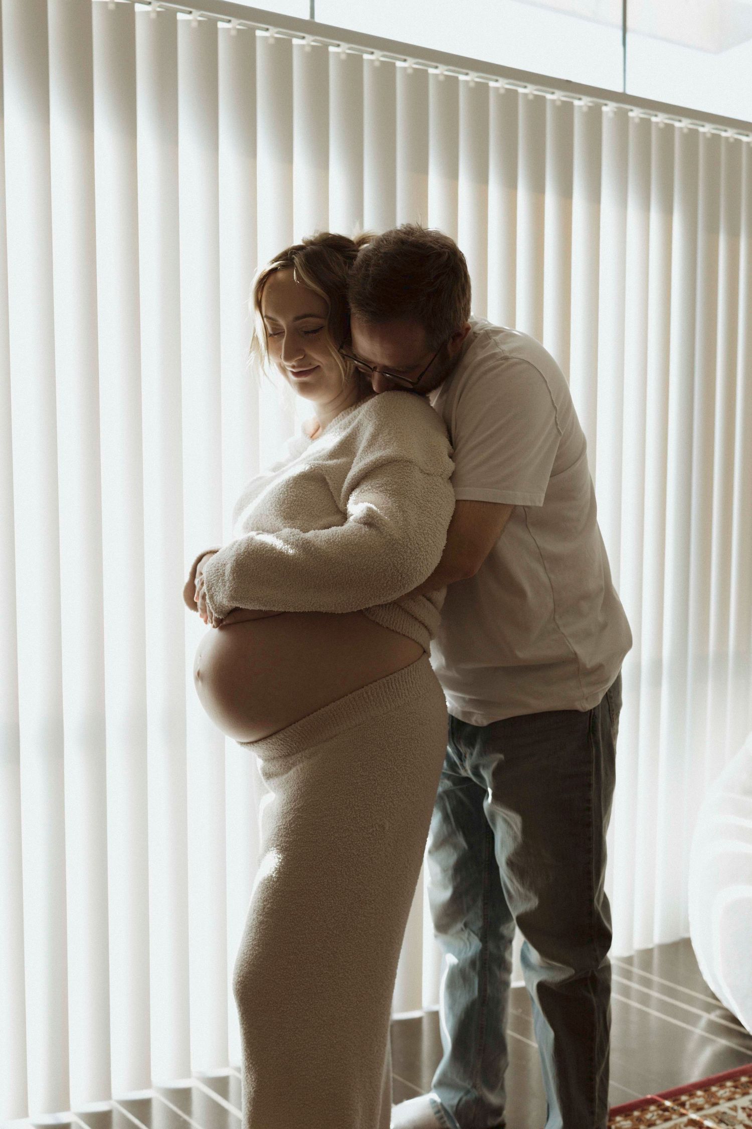 A couple hugging in a bright room with vertical blinds in the background, looking at a baby bump.