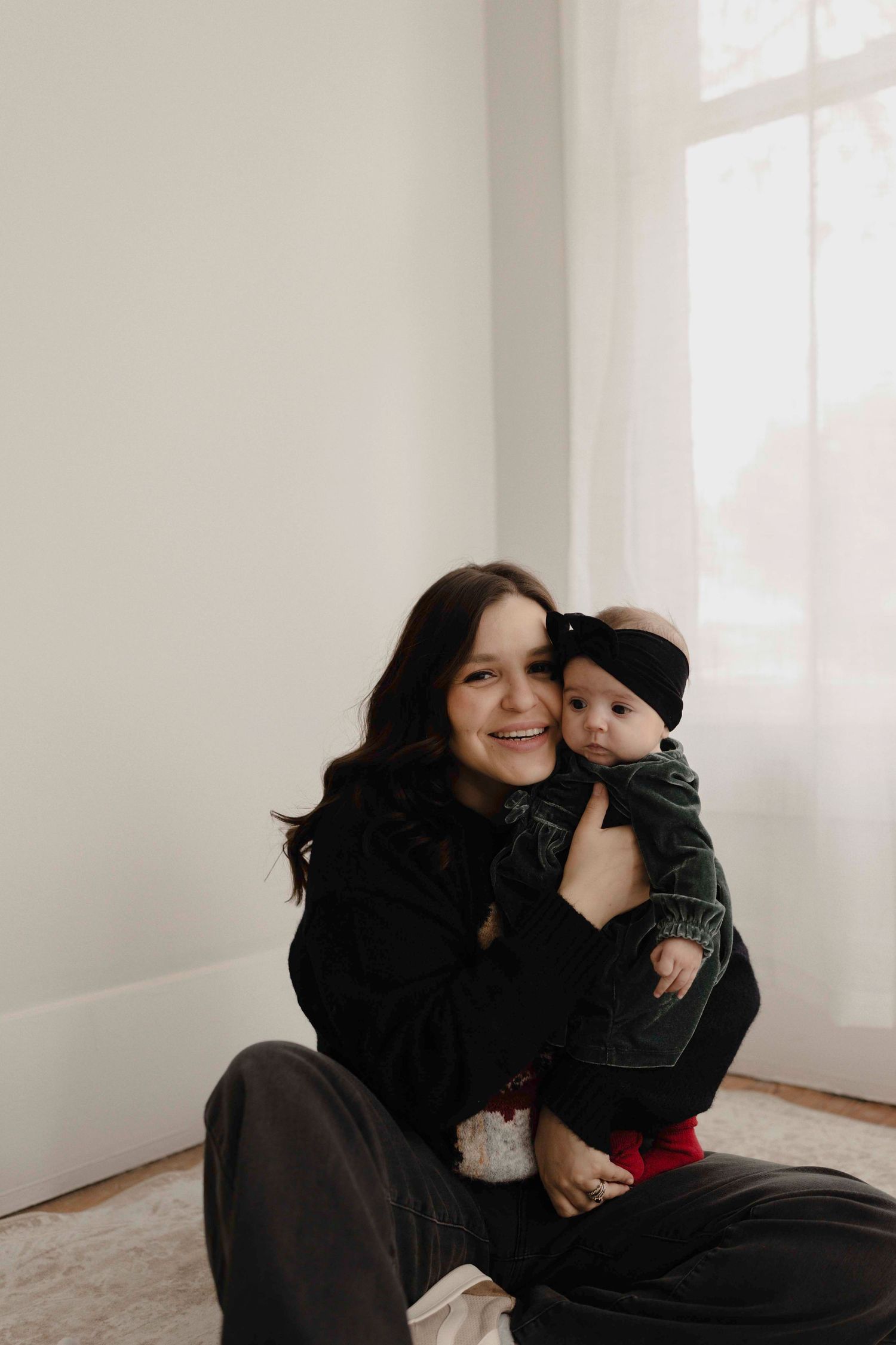 A mother and baby sit together by a bright window, sharing a tender moment in casual black clothing.