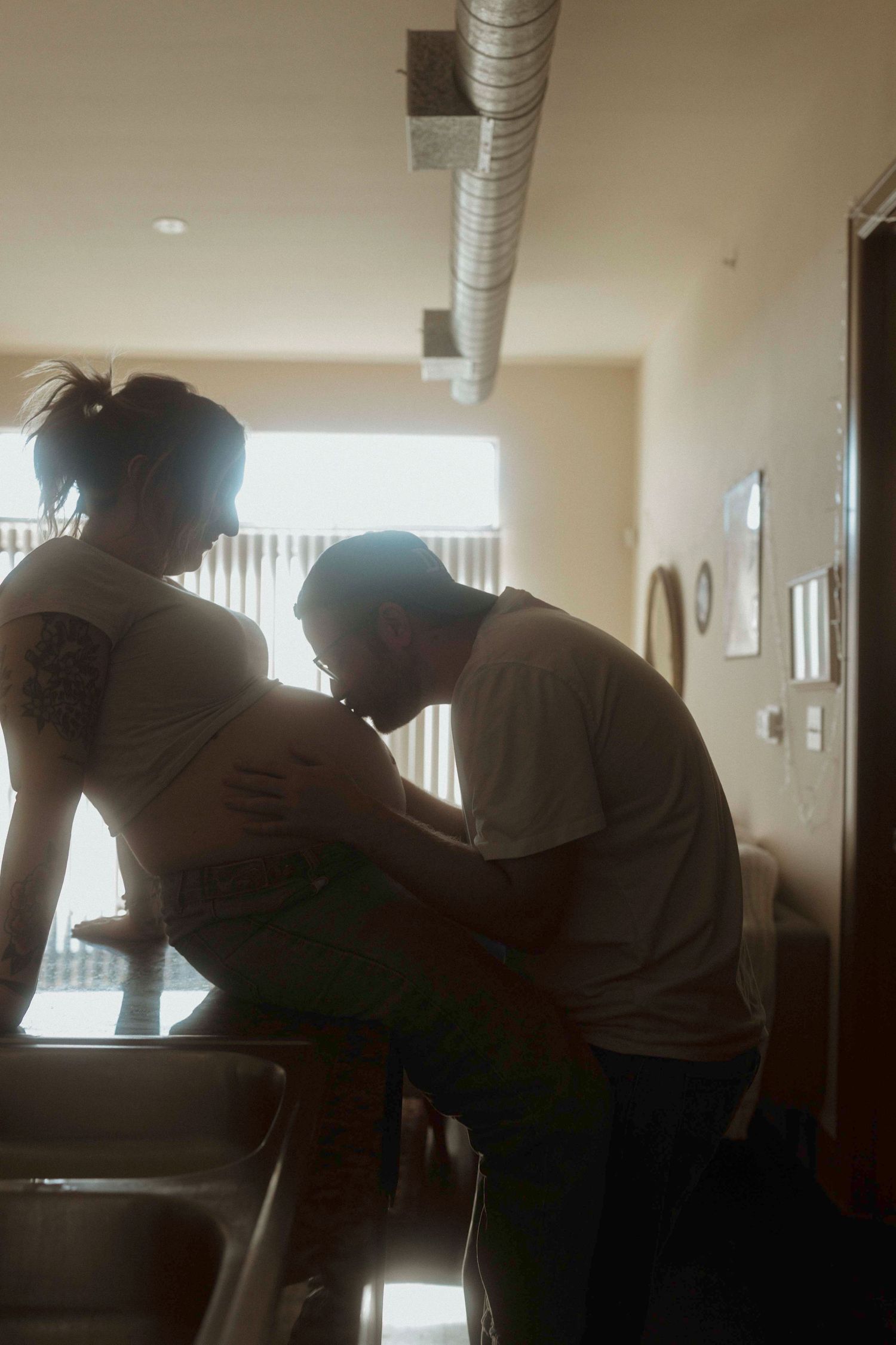 A silhouetted maternity couple shares an intimate moment in a sunlit indoor setting.