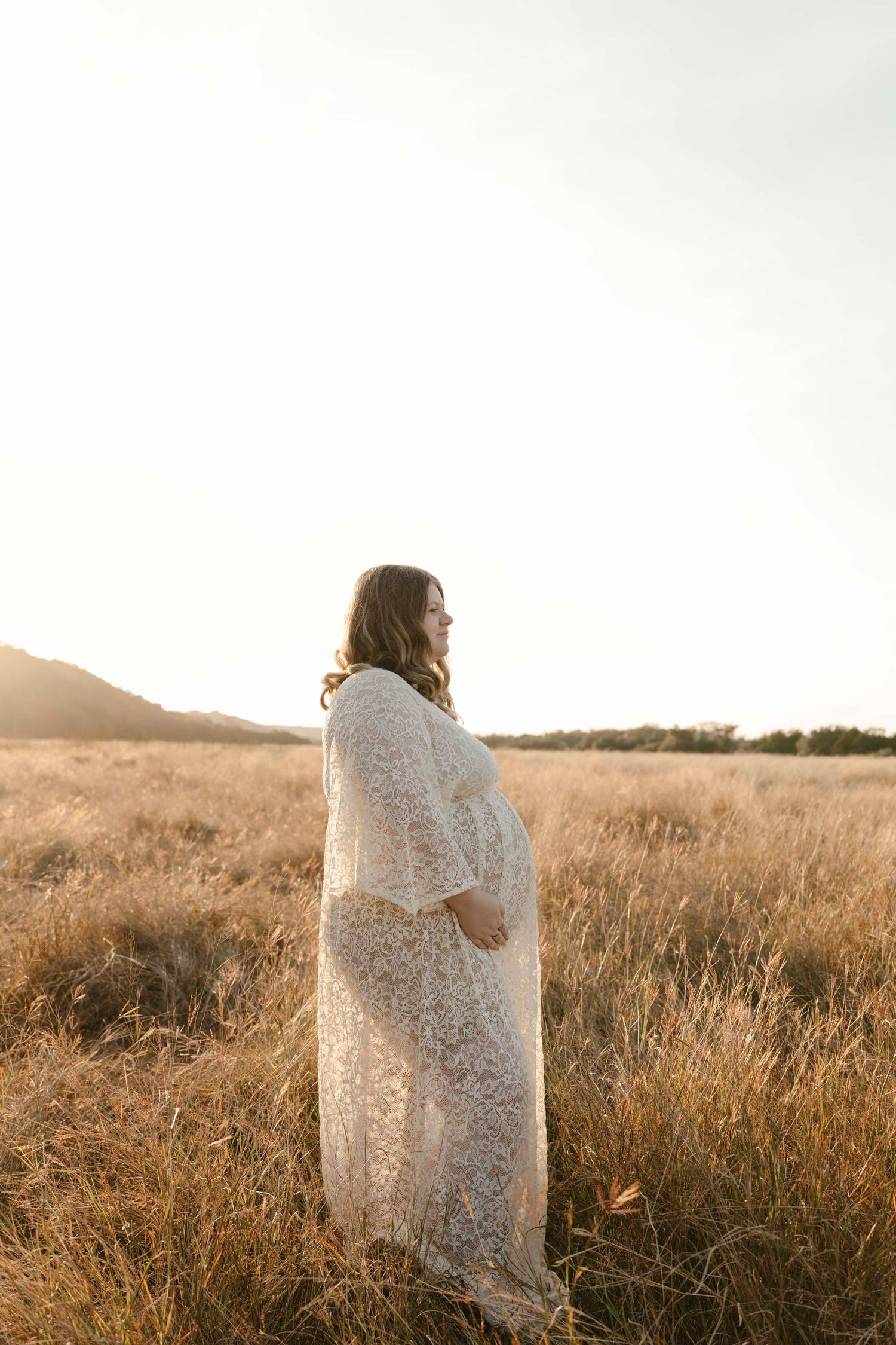 An expectant mother in a white lace dress stands in a golden field at sunset creating a dreamy silhouette.