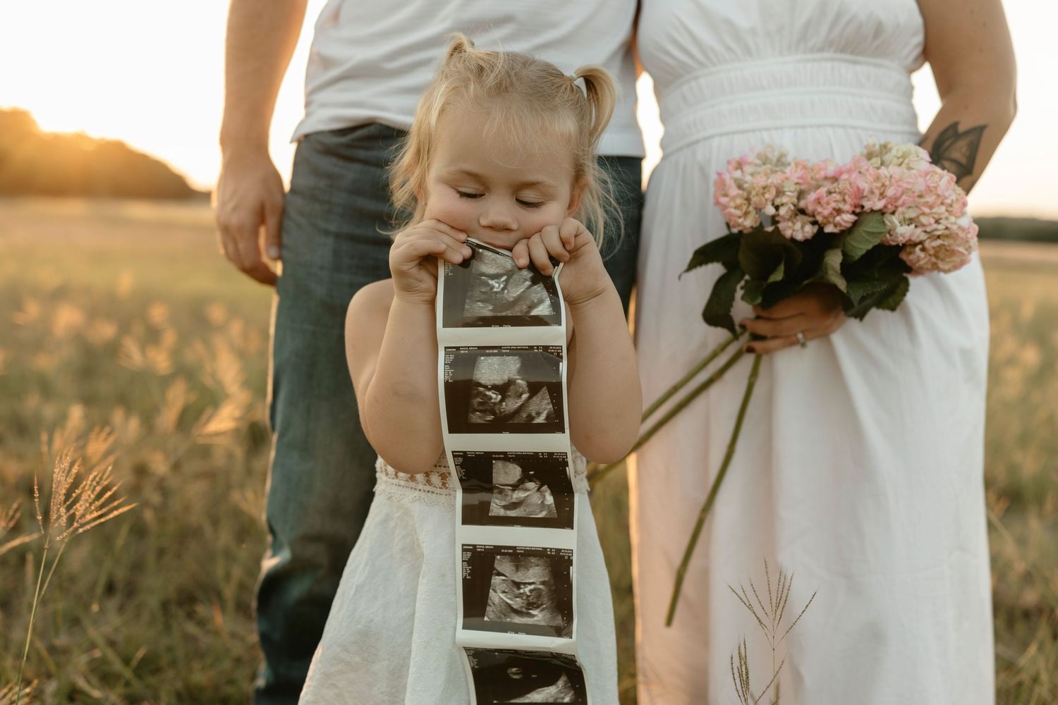 A small child in a white dress holds ultrasound photos while standing in a golden field at sunset with flowers nearby.