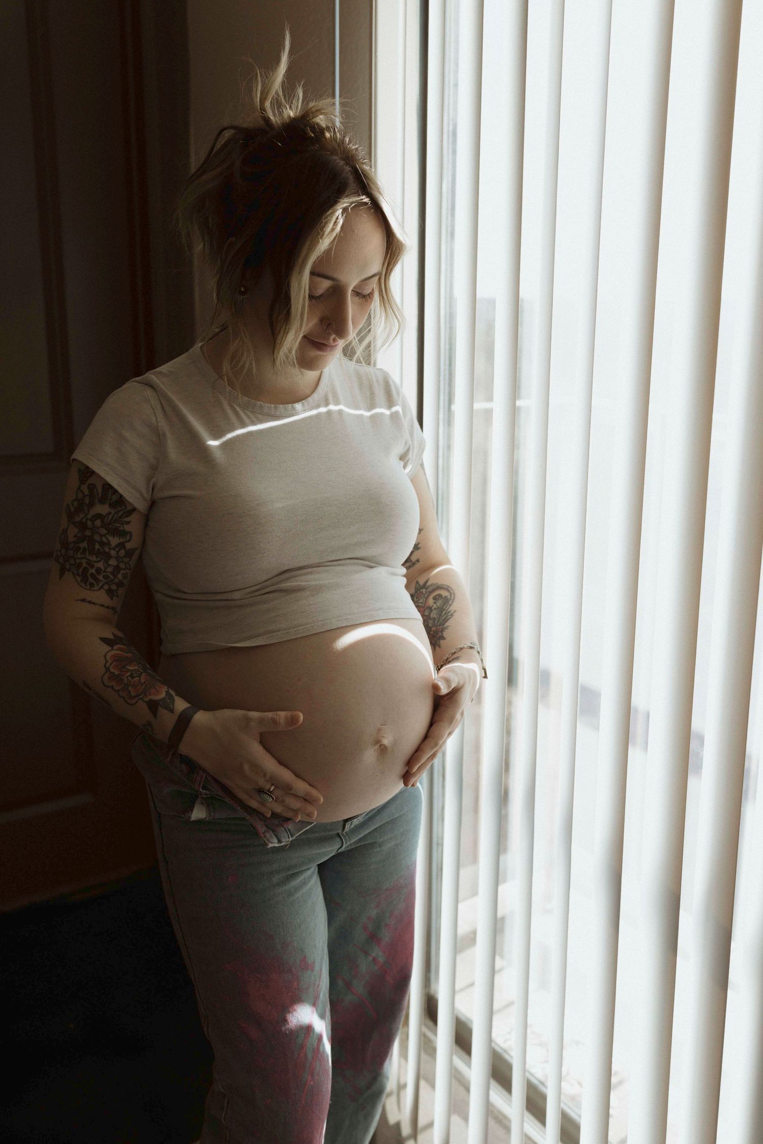 A pregnant person stands by vertical blinds in natural light showing their growing belly.