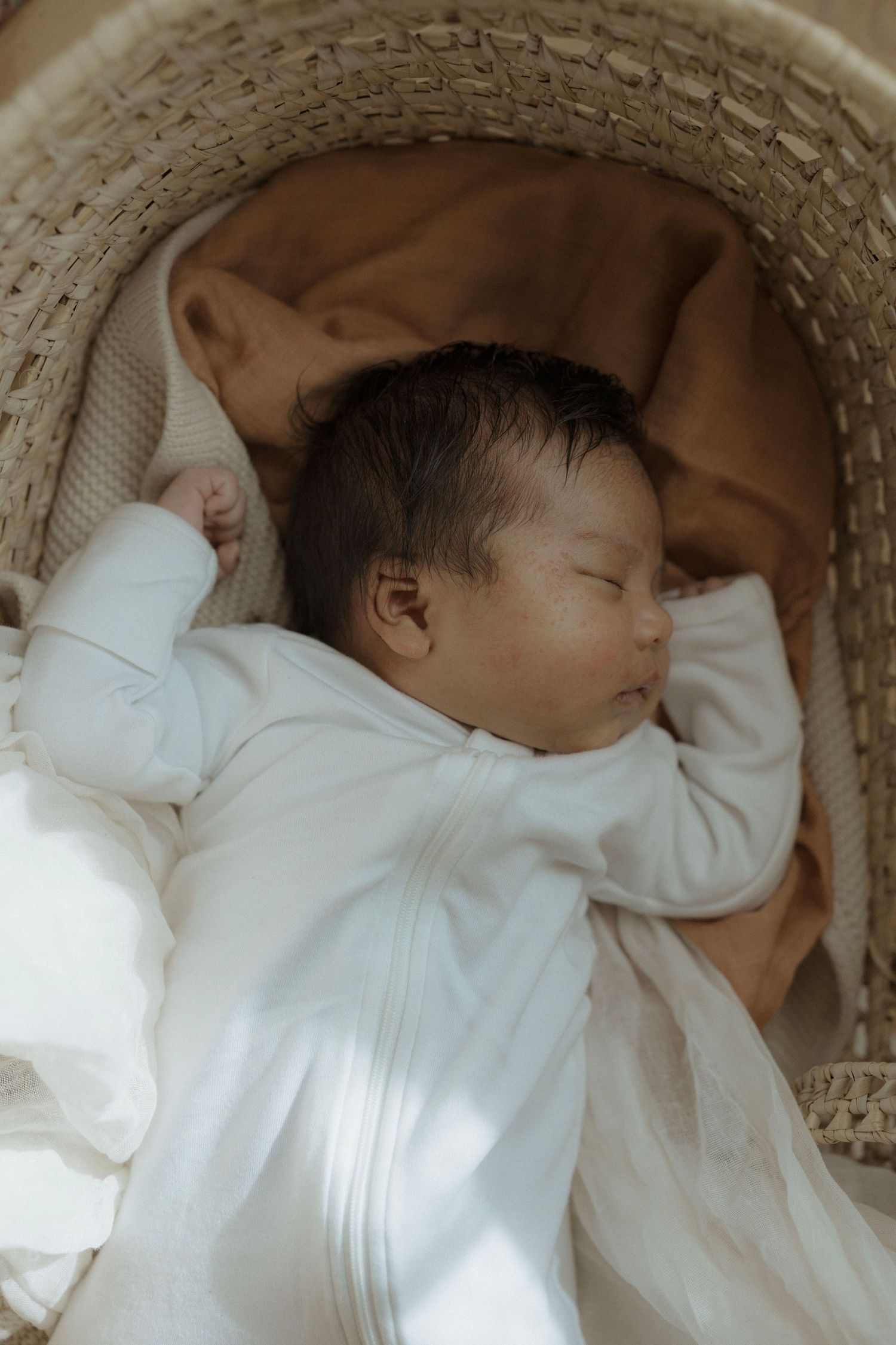 Newborn photo of peaceful sleeping baby in white clothing rests in cozy wicker bassinet with tan blanket.