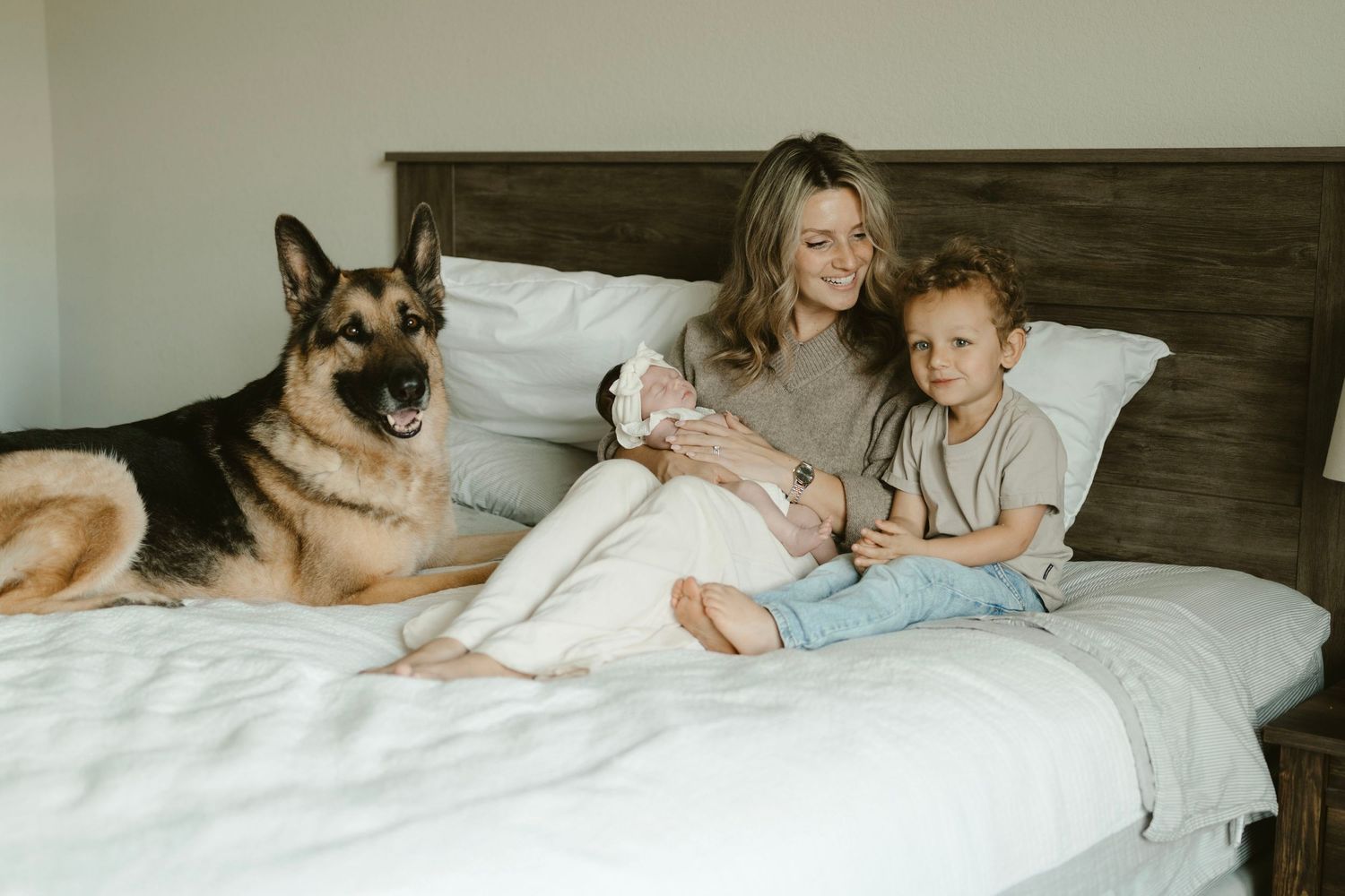 A German Shepherd dog rests on a bed with people sitting against a wooden headboard.