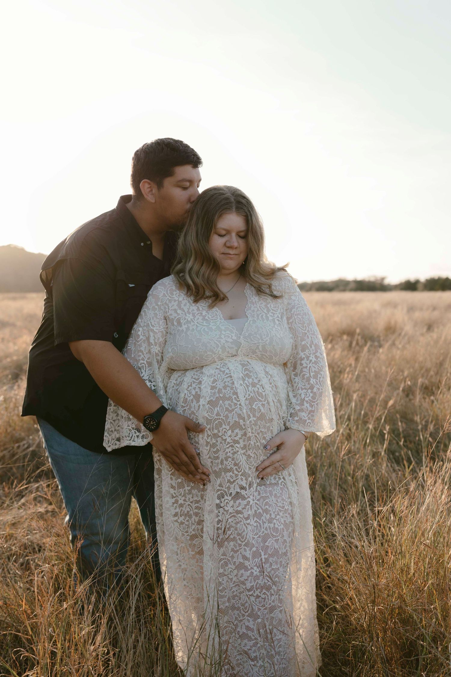 A couple shares an intimate moment in a golden field during sunset, wearing casual and white lace attire.