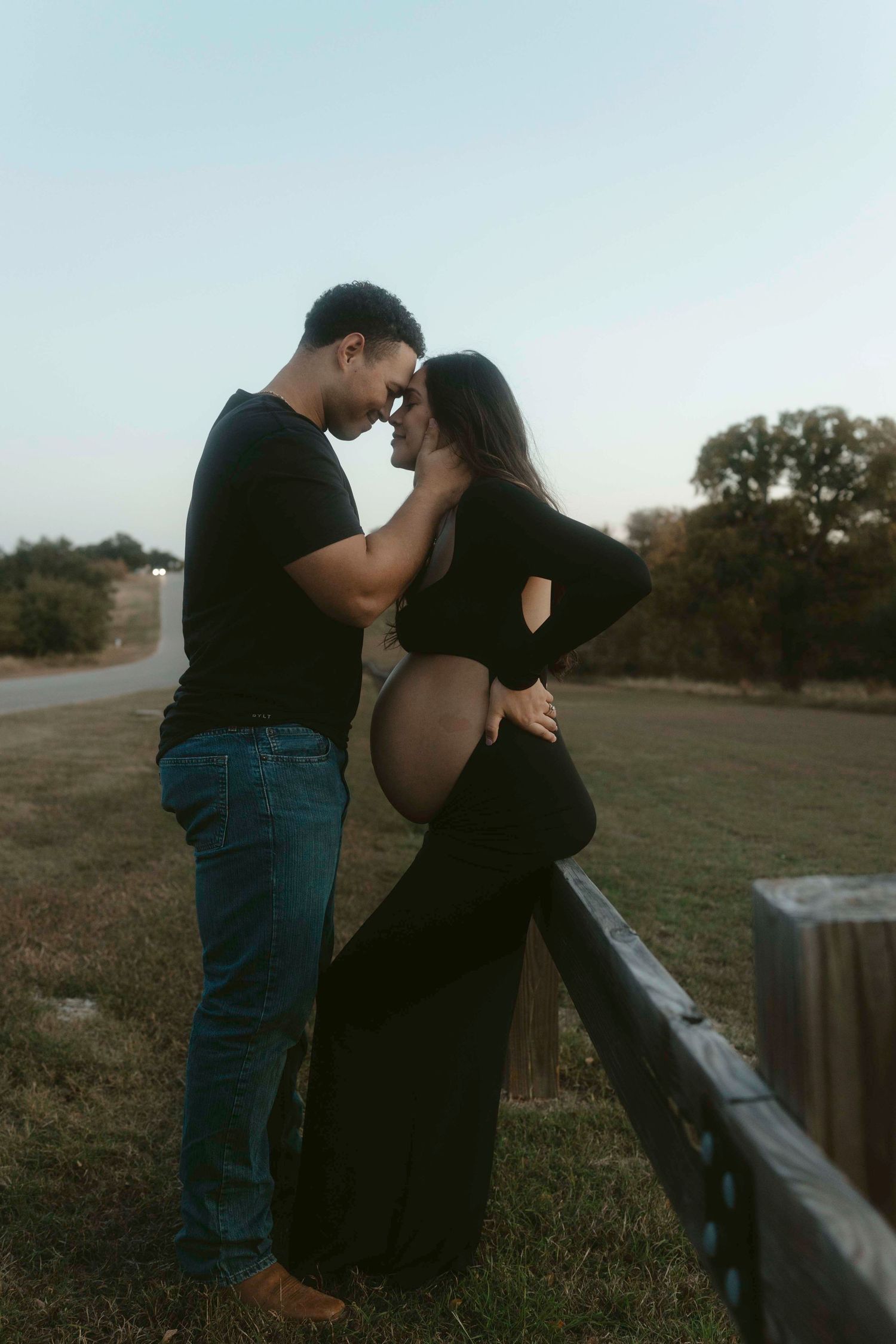 A couple shares an intimate moment during a sunset maternity photoshoot along a rustic fence.