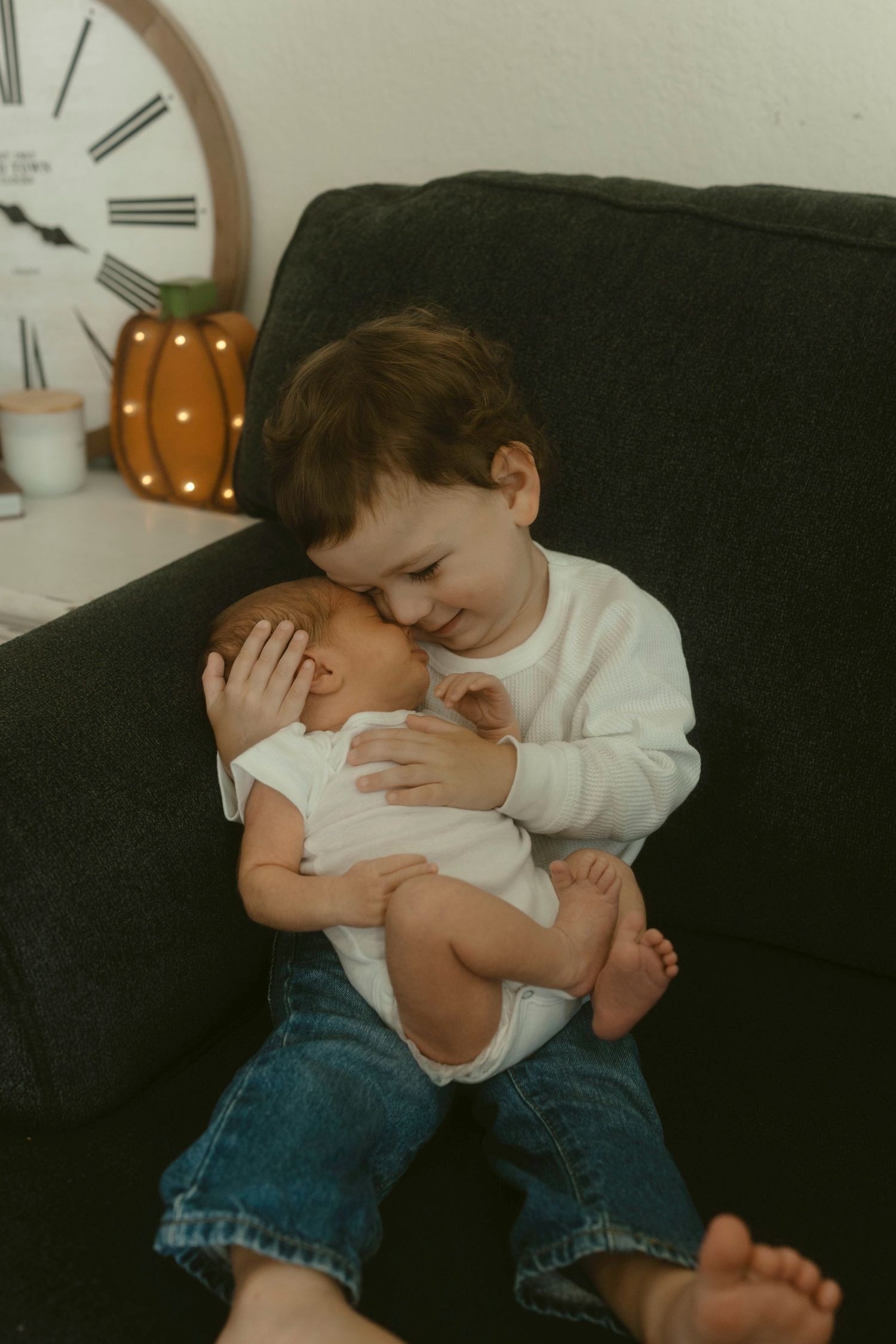 A toddler wearing a white sweater and jeans sits on a dark couch while holding a baby in their arms.