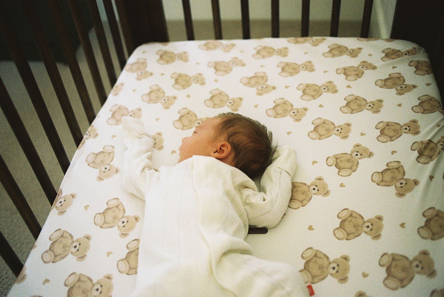 Baby sleeping peacefully in white crib with teddy bear patterned sheets.