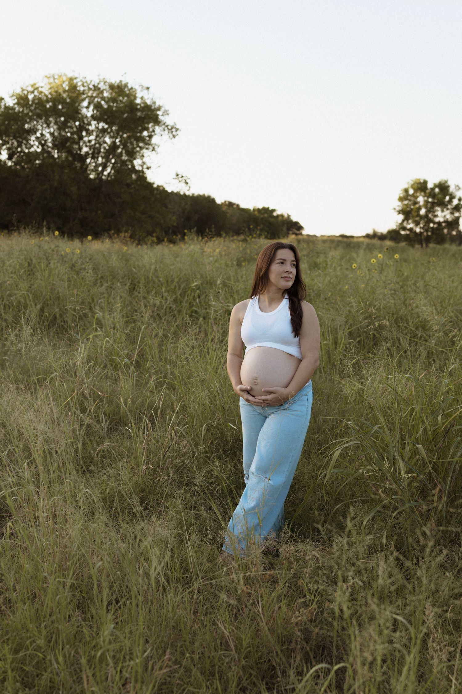 Maternity photoshoot in a rustic field at sunset shows mother-to-be wearing light blue dress and white top.