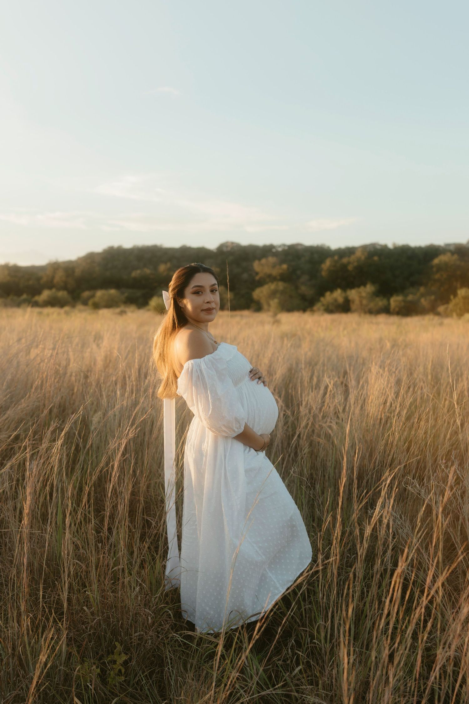 A pregnant woman in a flowing white dress twirls gracefully through a sun-lit field of tall grass at golden hour.