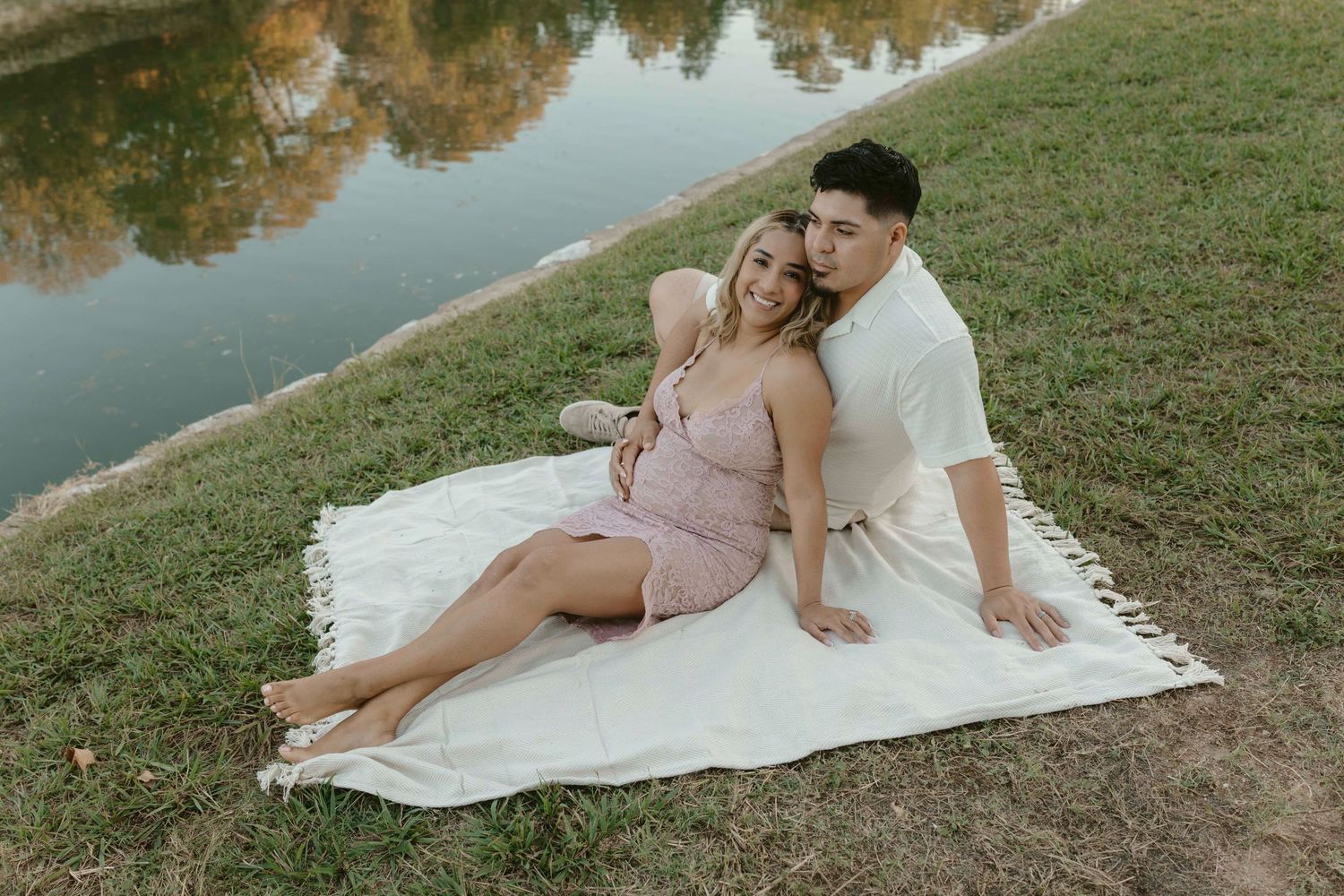 A couple in light-colored outfits reclines on a white blanket near a lake during a romantic outdoor photoshoot.