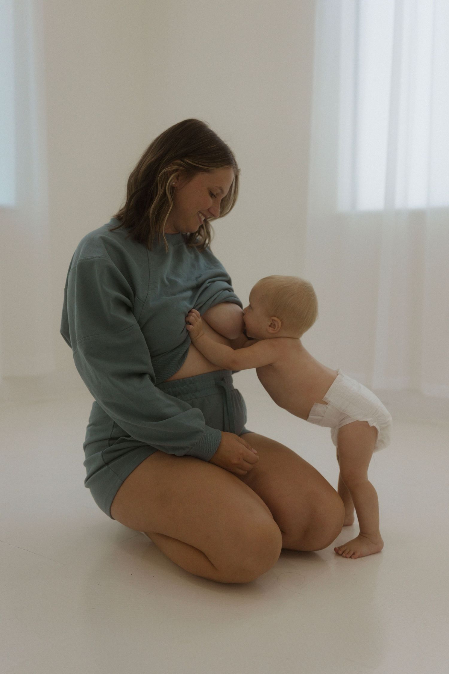 A mom in a blue lounge set sits on the floor tenderly breastfeeding and bonding with their baby in a light-filled room.