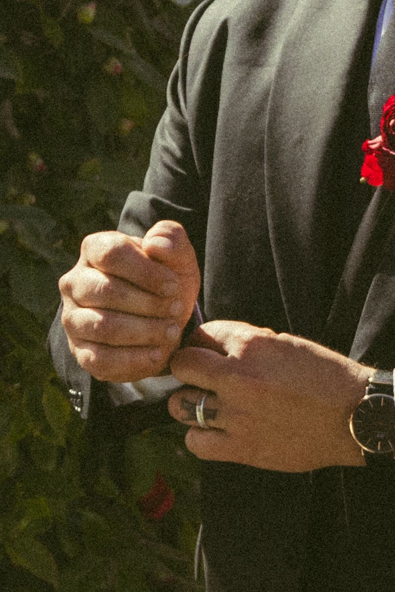 Man in dark suit adjusting cufflinks, wearing a watch and ring, with red boutonniere, outdoors.