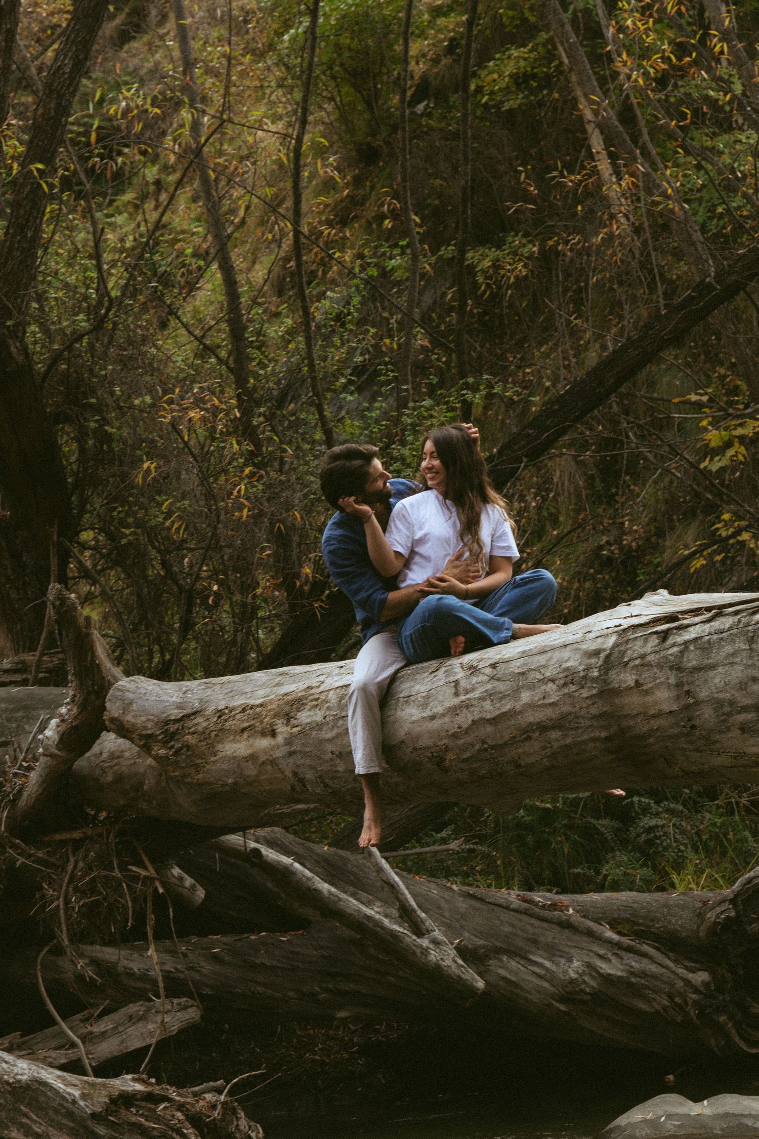 Couple sitting on a fallen log in a lush forest, smiling at each other in a romantic outdoor setting.