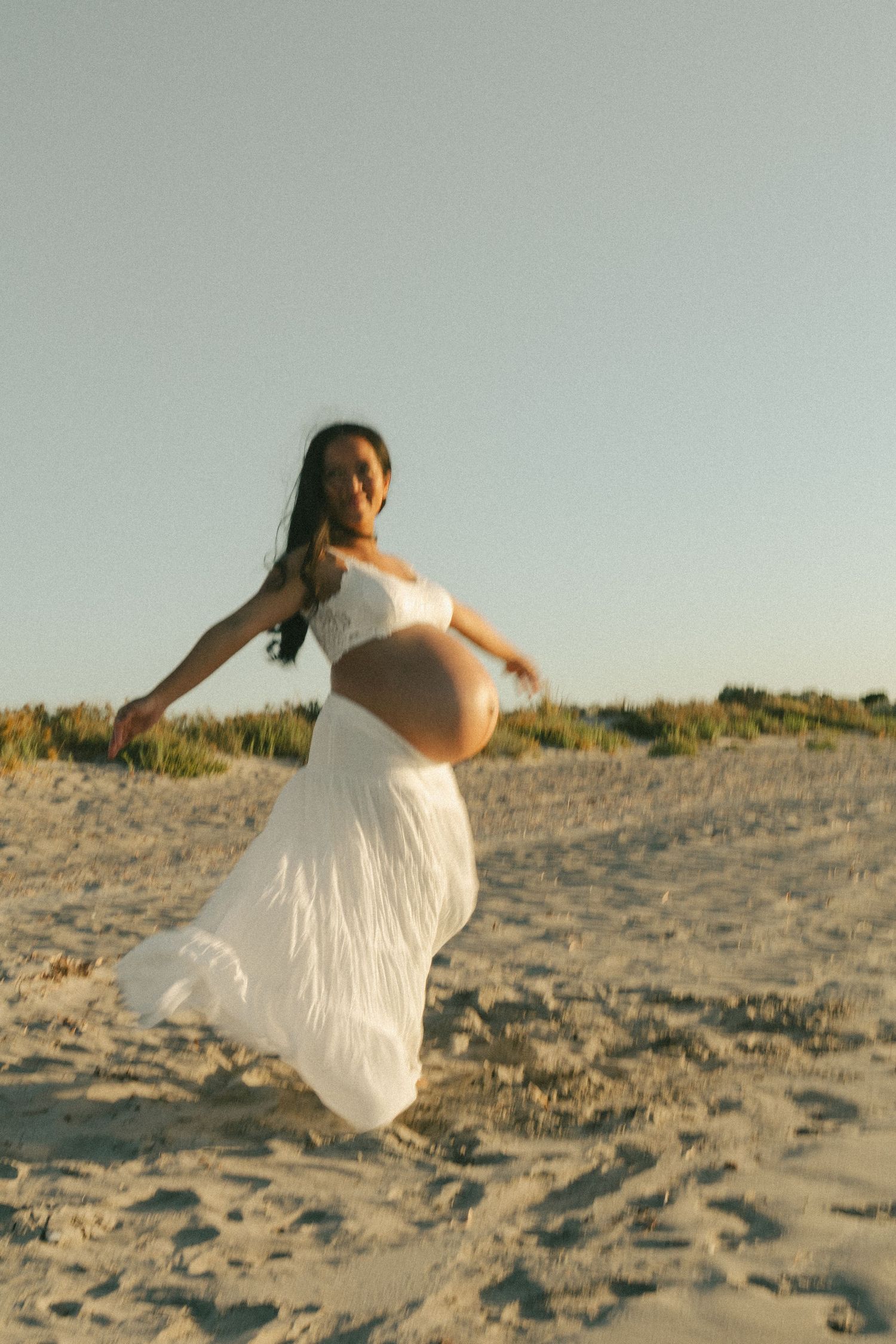 Pregnant woman in white crop top and flowing skirt poses gracefully on sandy beach dunes at golden hour.