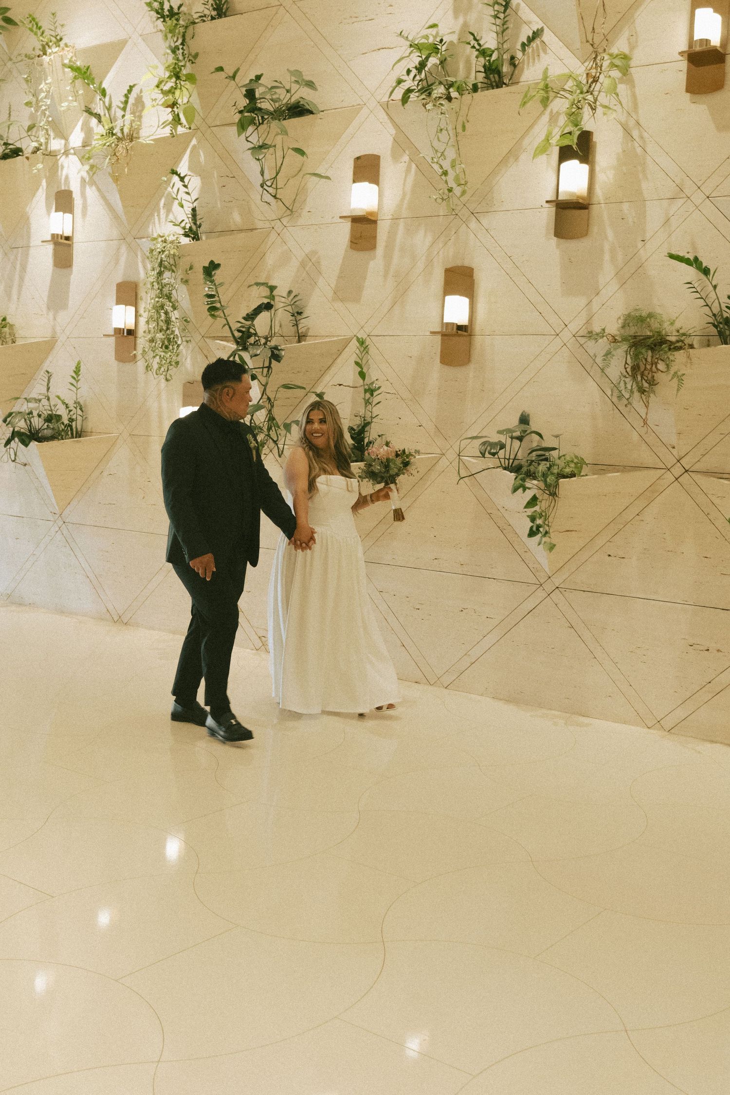 Bride and groom holding hands on dance floor before geometric plant wall with warm lighting.