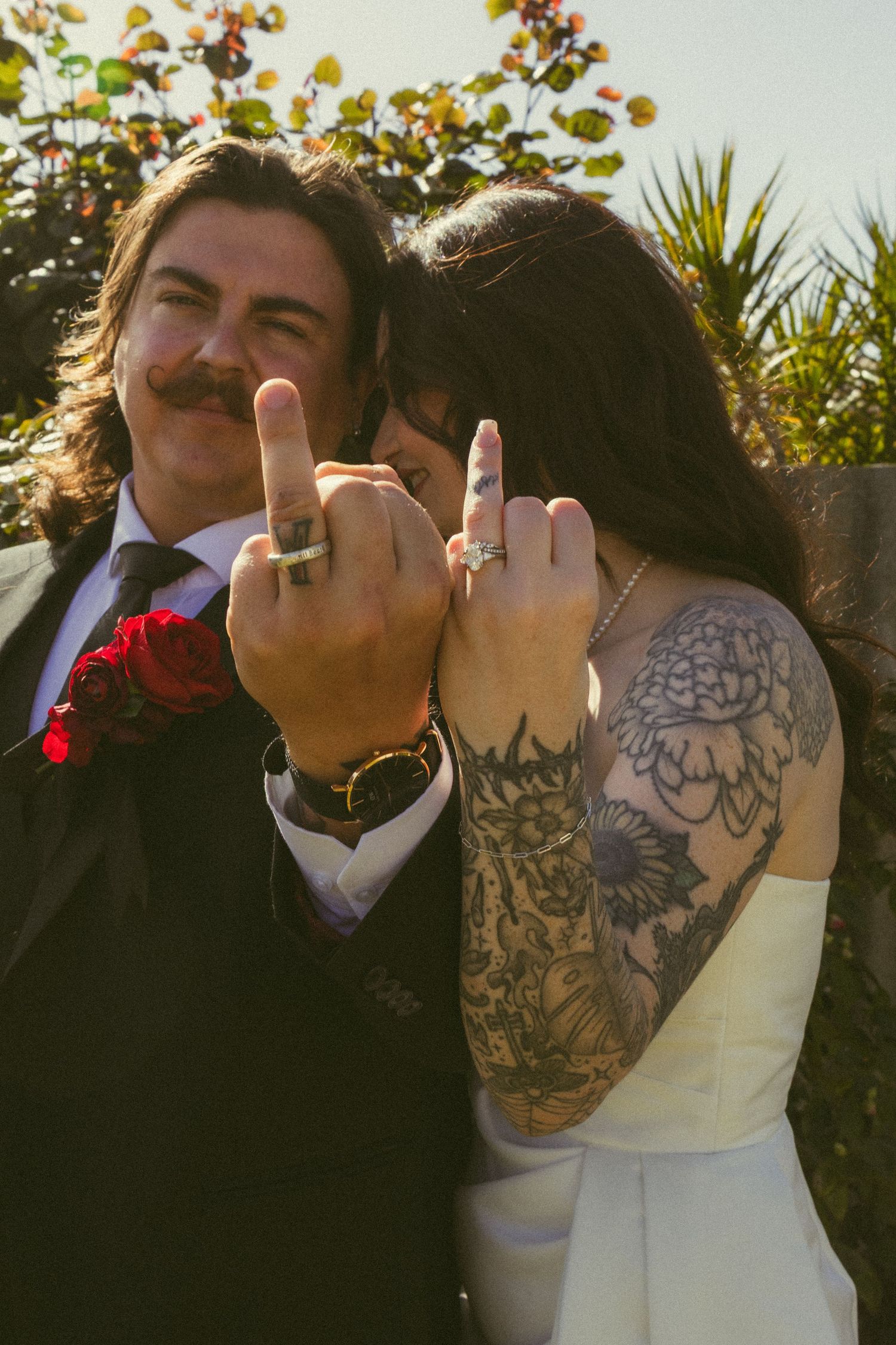 Newlywed couple showing off wedding rings with tattooed bride in white dress and groom in suit with red boutonniere.