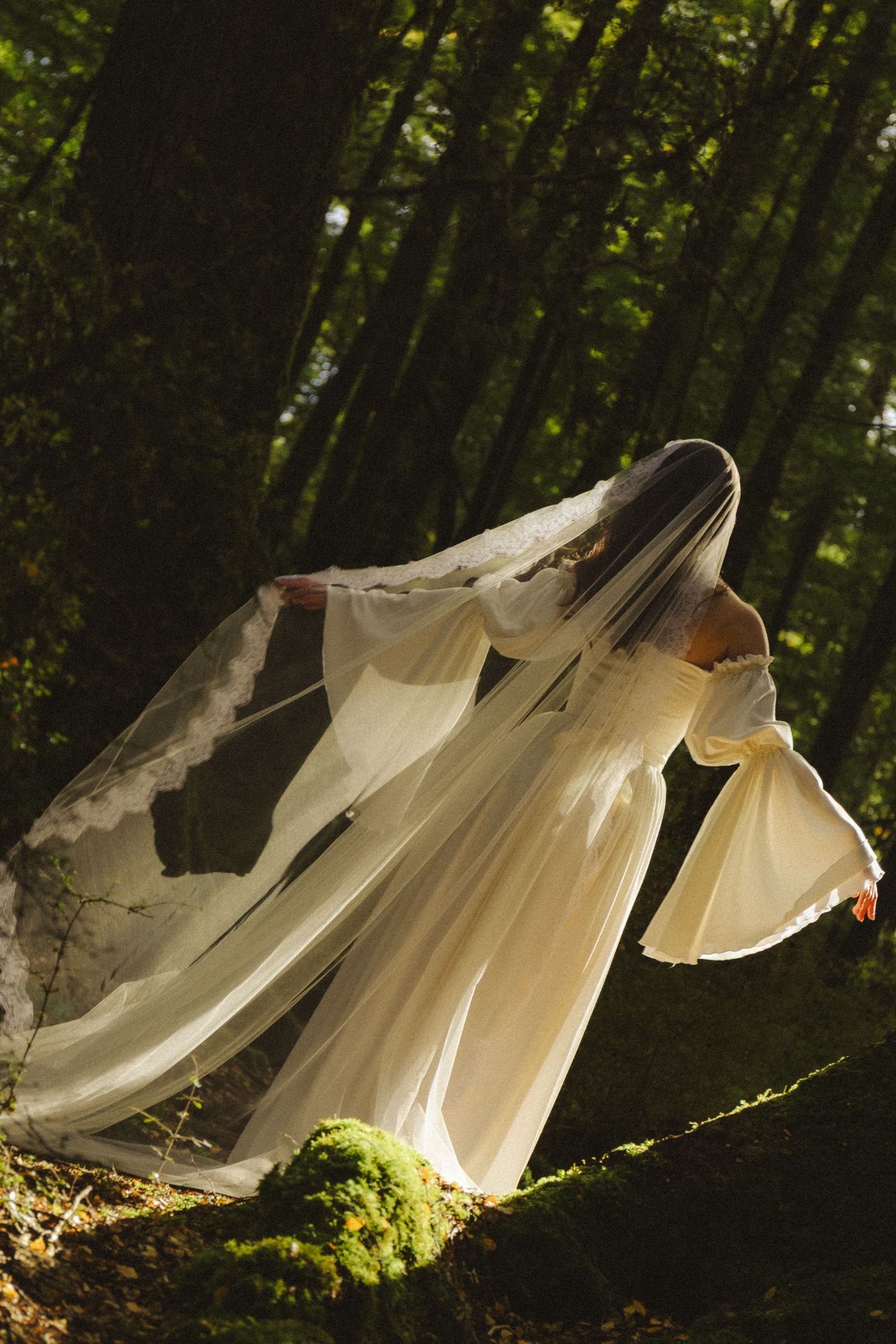 A bride in a flowing white gown and lace veil stands gracefully among mossy trees in a sunlit forest.