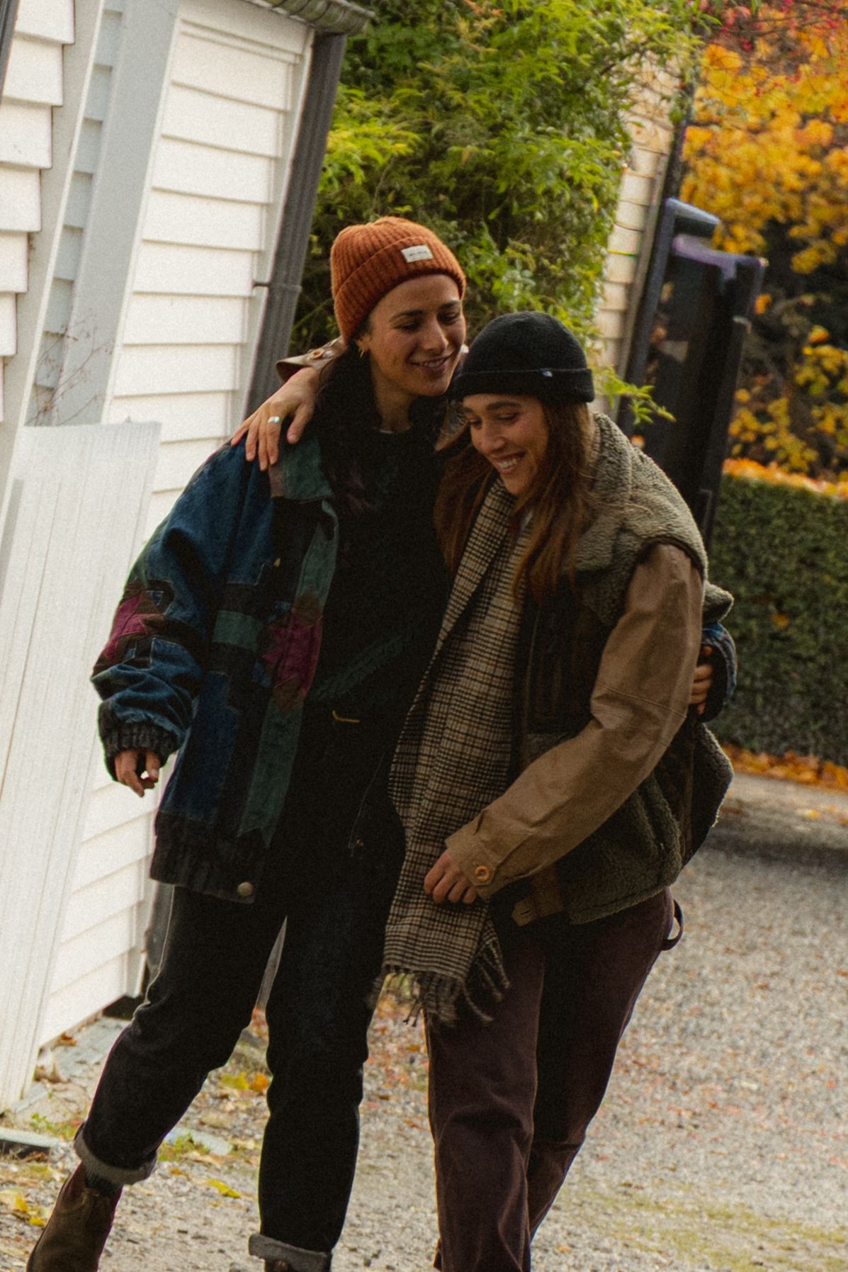Two friends in winter hats and layered jackets walk together arm in arm on a gravel path during autumn.