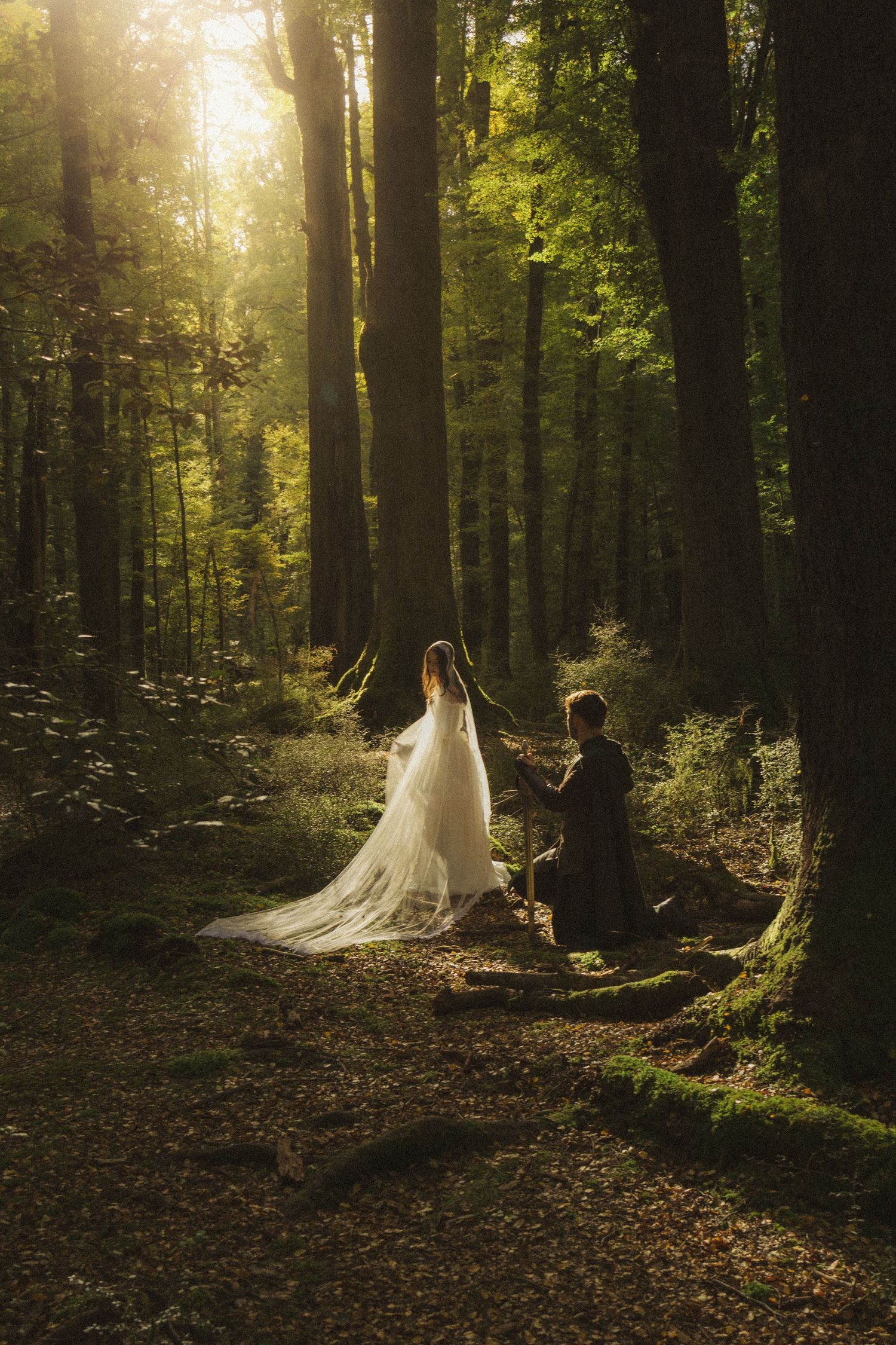 Groom kneels before bride in sunlit forest during romantic outdoor wedding.