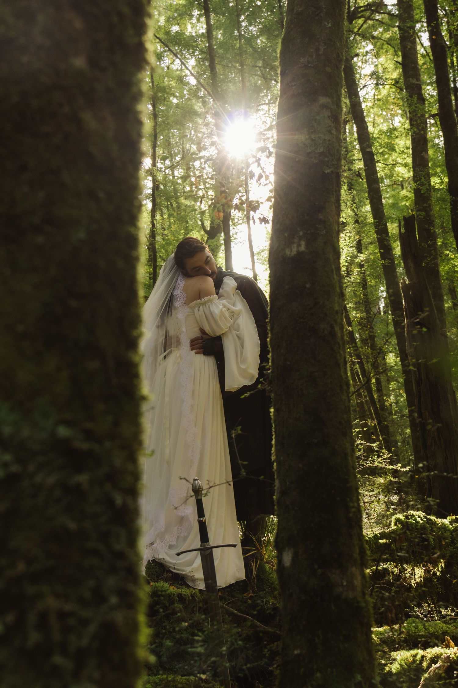 A bride and groom embrace in a sunlit forest, the bride in a white gown and veil, the groom in dark medieval attire with a sword.
