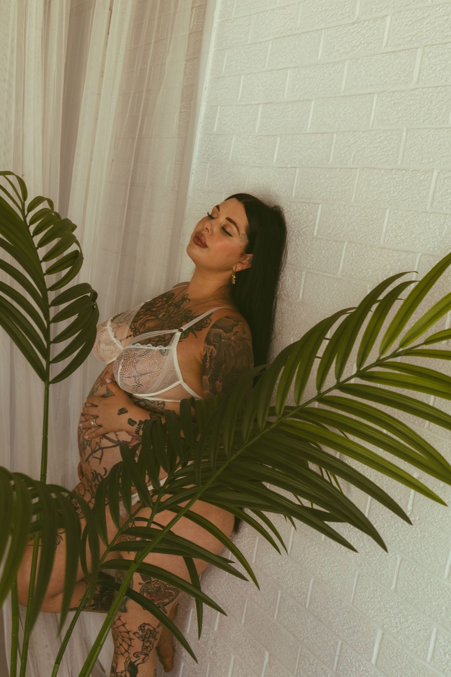 Tattooed woman in white lingerie posing against brick wall with tropical palm leaves in foreground.