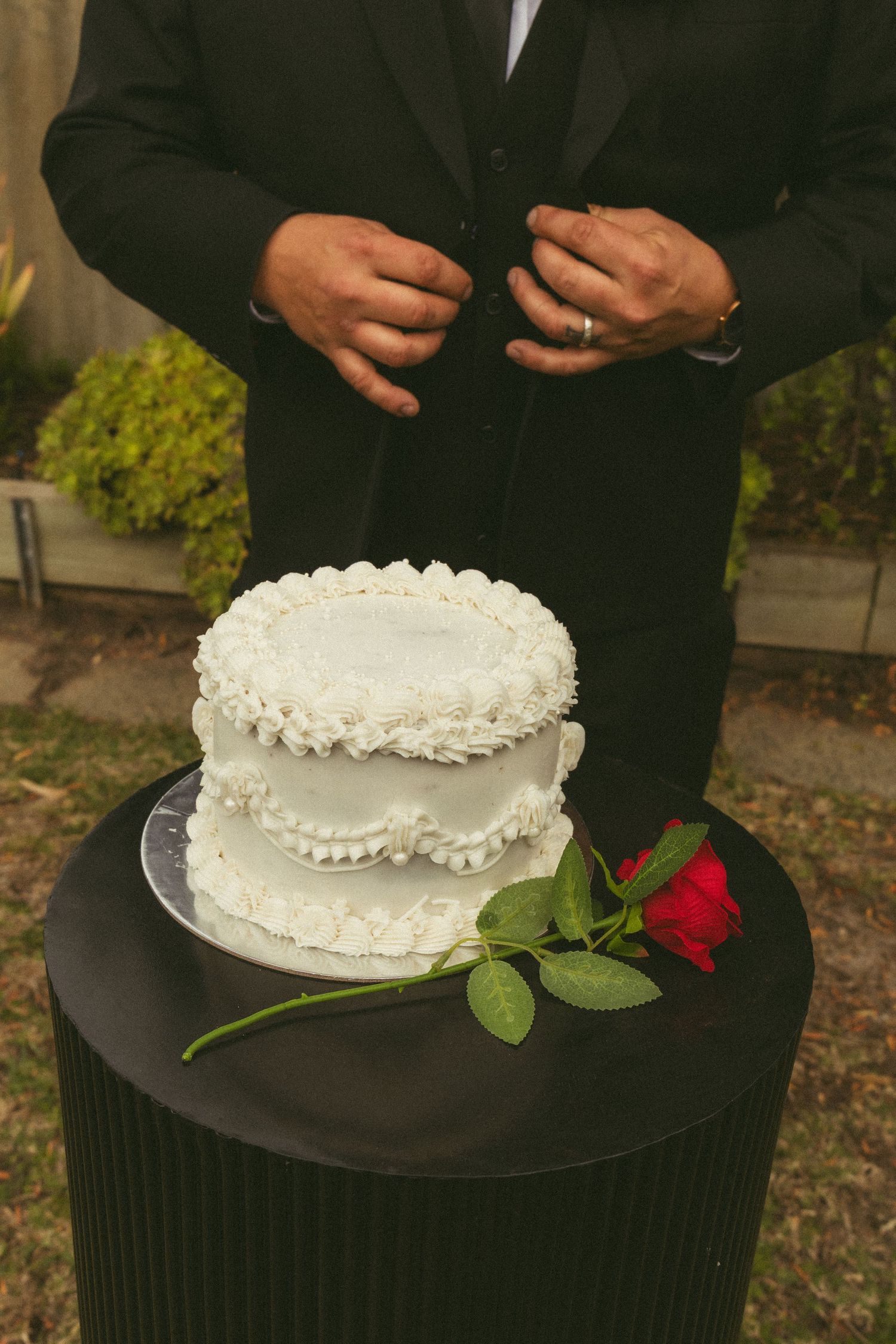 Two-tier white wedding cake with red rose on black table, man in black suit standing behind it outdoors.