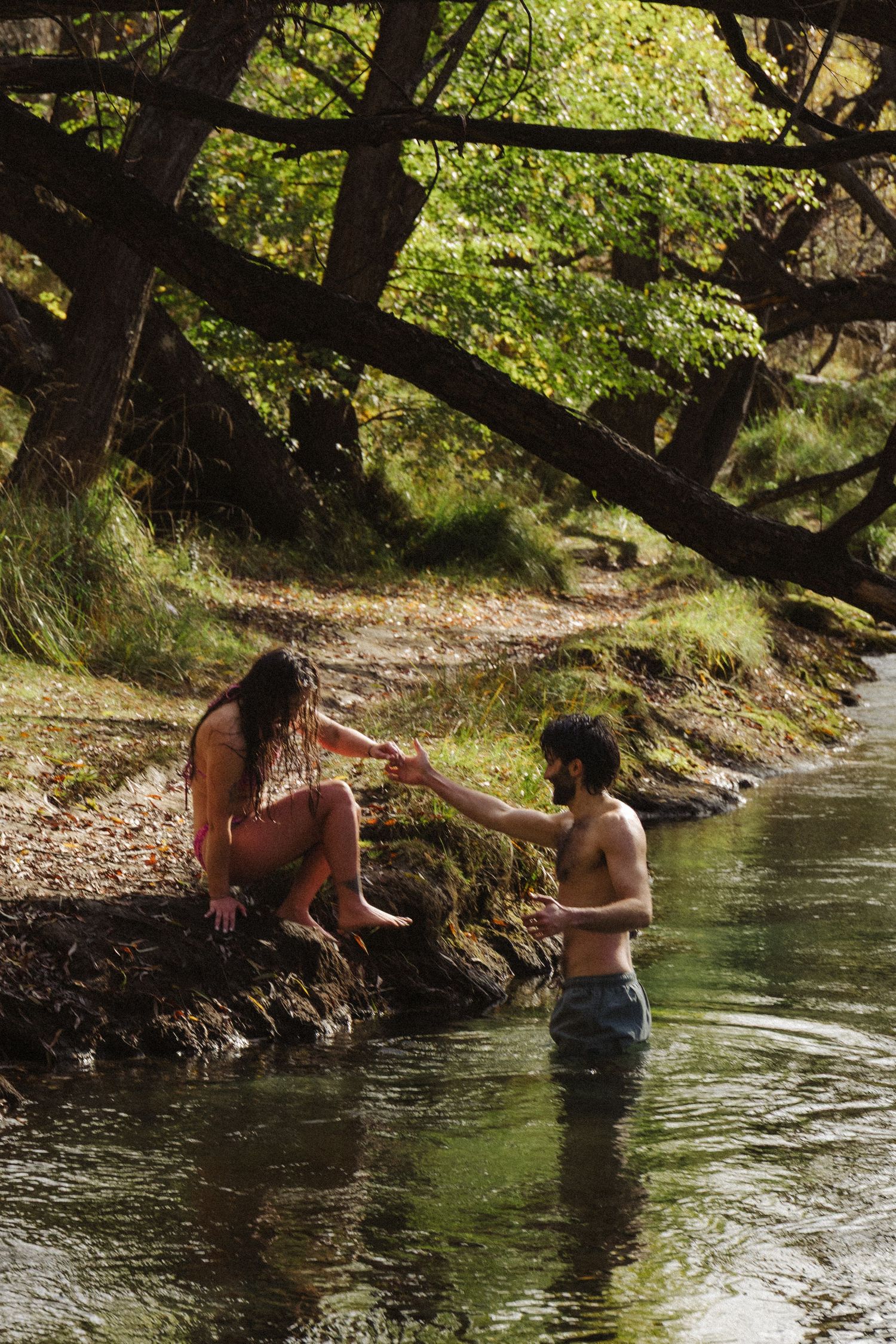 Couple holding hands by a forest stream, woman sitting on rocks in bikini, man standing in water shirtless.