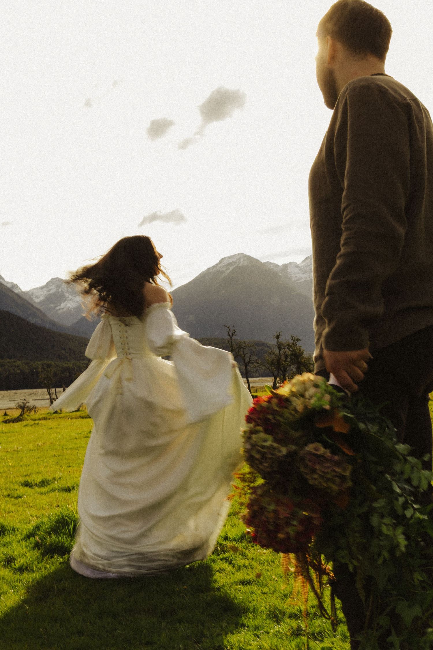 Bride in bell-sleeve gown runs toward mountains as groom holds bouquet.