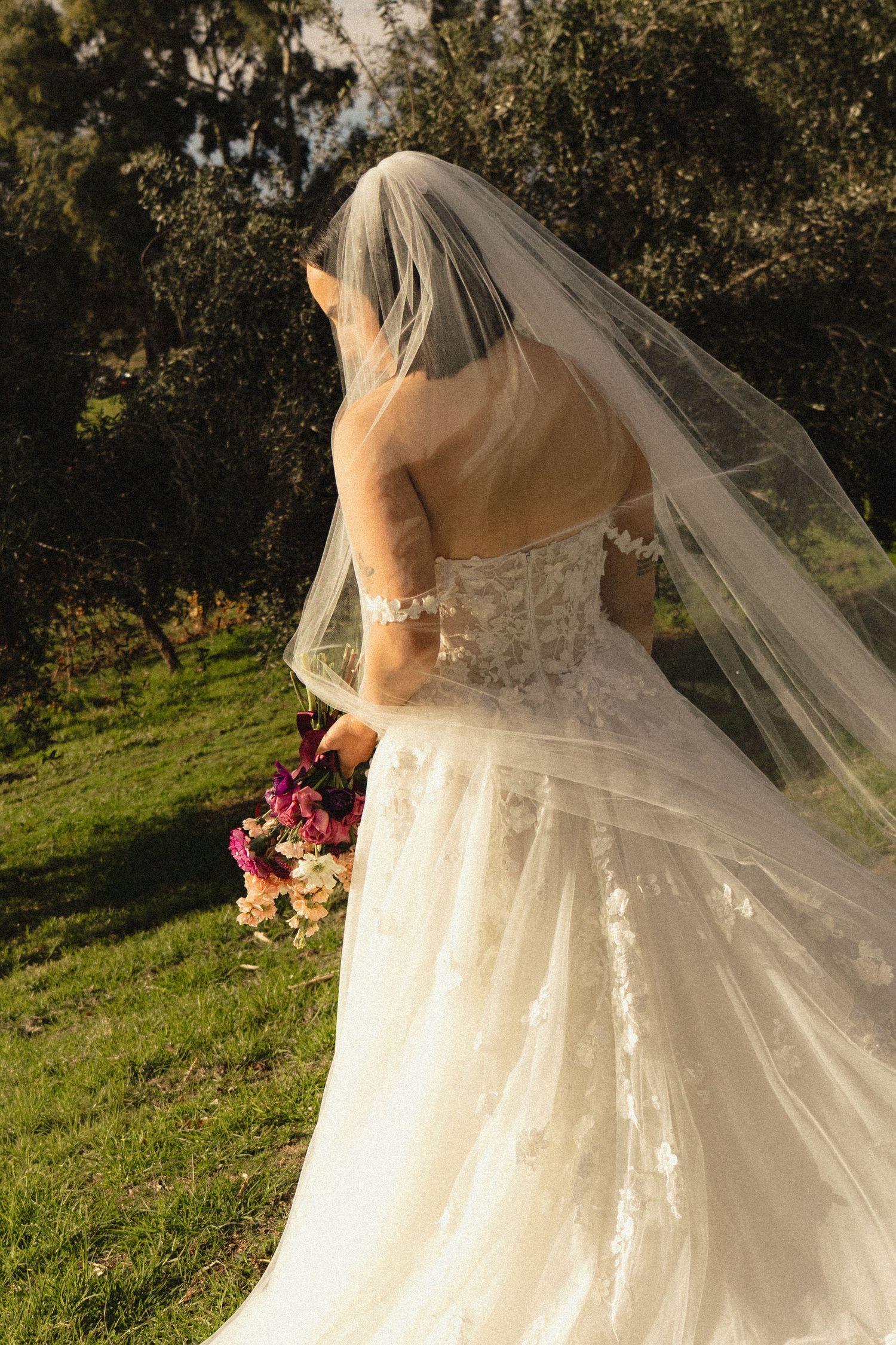 Bride in white lace gown and veil holding colorful bouquet, standing in lush green garden, viewed from behind.
