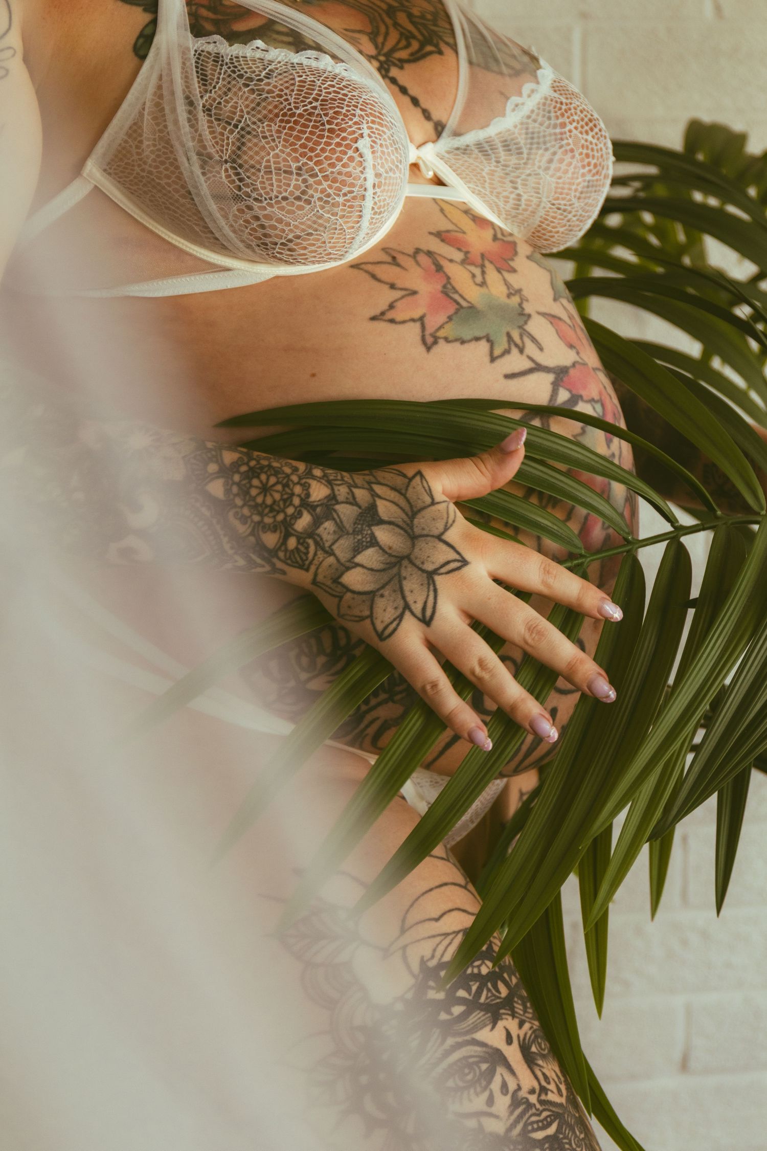 Tattooed woman in white lace lingerie posing with tropical palm leaves, showcasing floral and mandala body art.