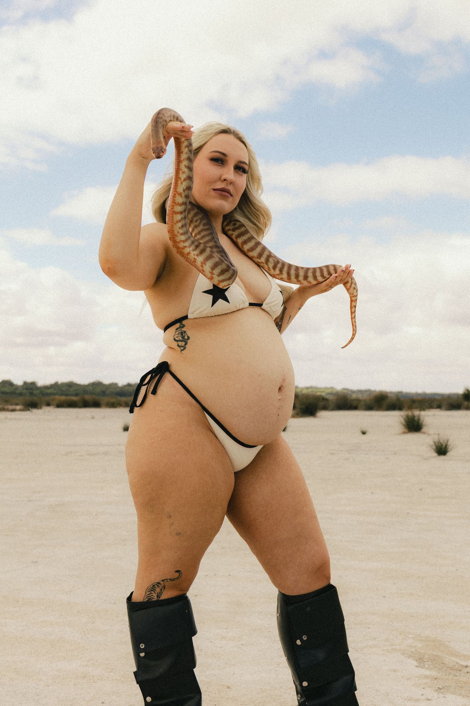 Pregnant woman in white star bikini and black boots holding a snake in a desert landscape.