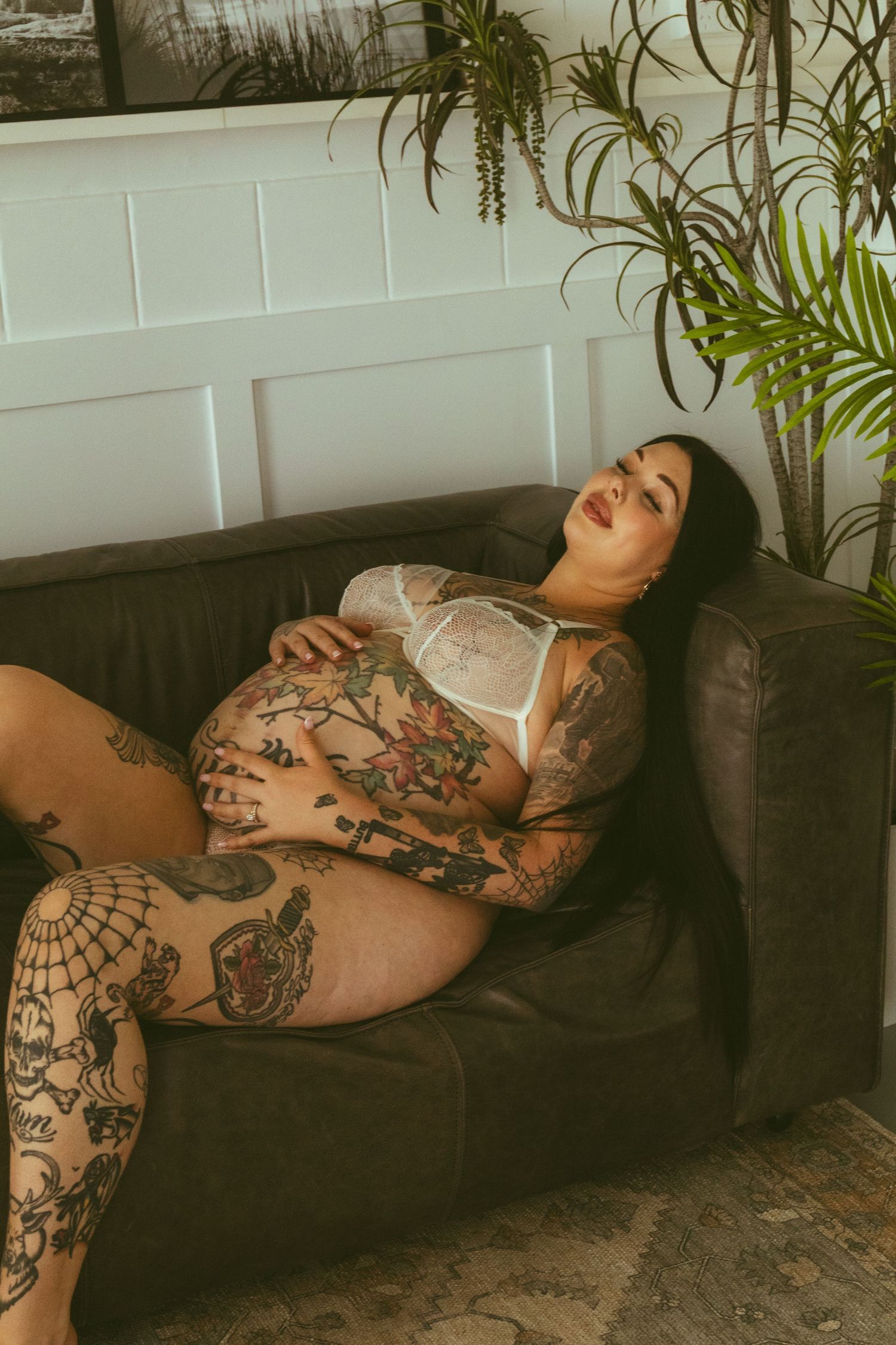 Heavily tattooed pregnant woman in white lace lingerie relaxing on a dark sofa near indoor plants.