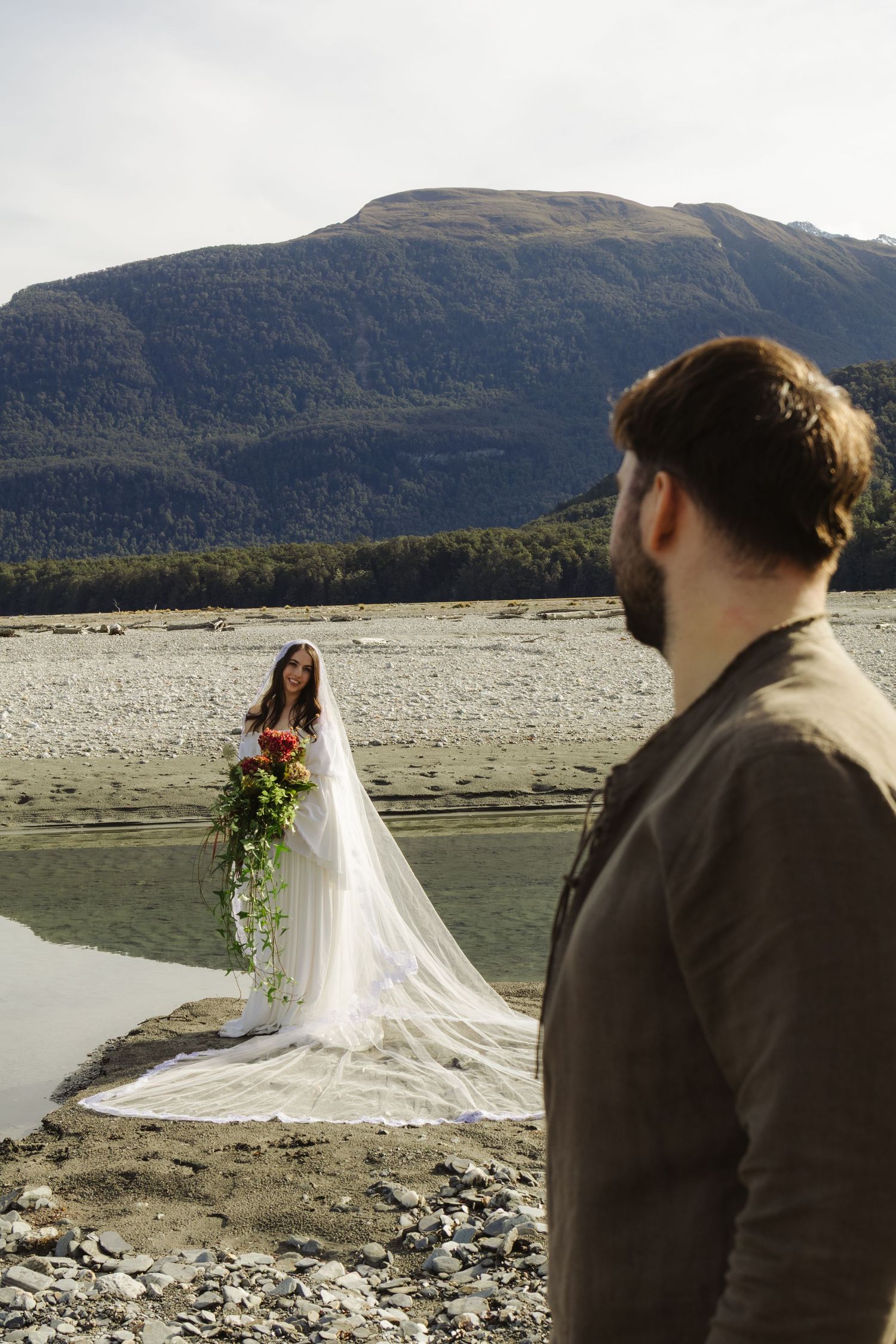 Bride in white gown with floral bouquet stands by mountain lake as groom watches in foreground.