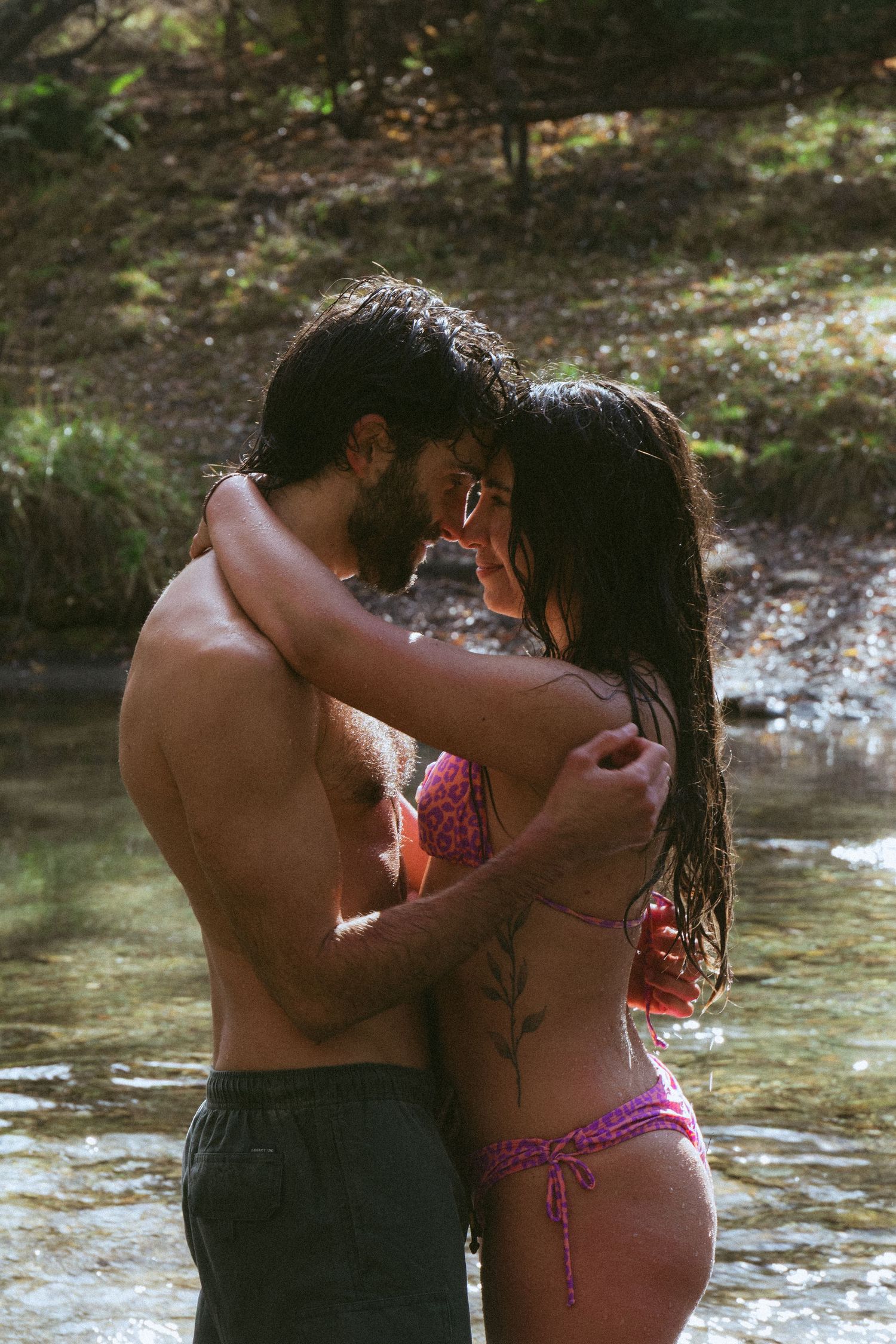 Romantic couple embracing in a shallow forest stream, woman in pink bikini with floral tattoo, man shirtless.