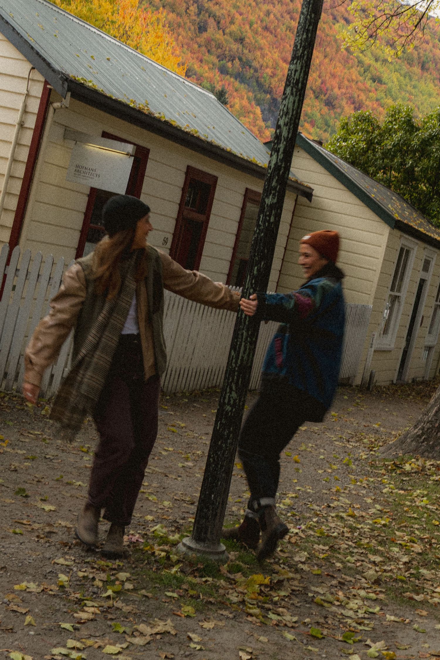 Two people in autumn outfits hold a tree pole outside white buildings with colorful fall foliage hillside backdrop.