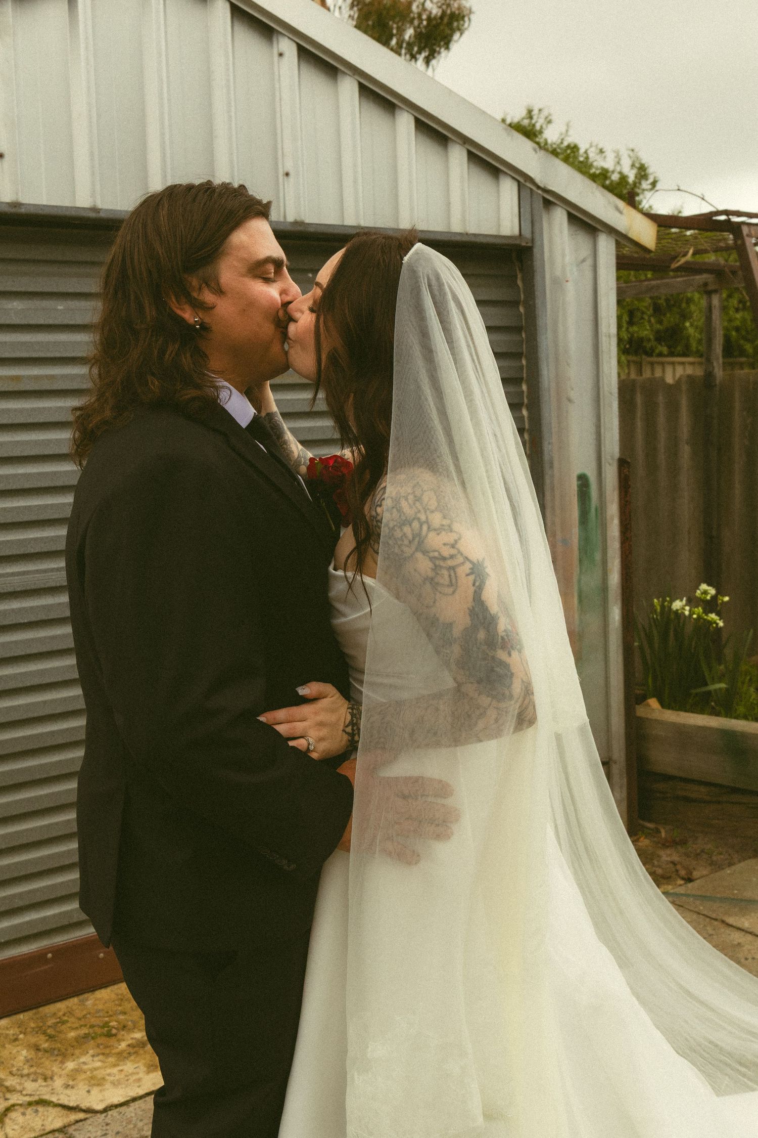 Tattooed bride in white gown and veil kissing long-haired groom in black suit outdoors.