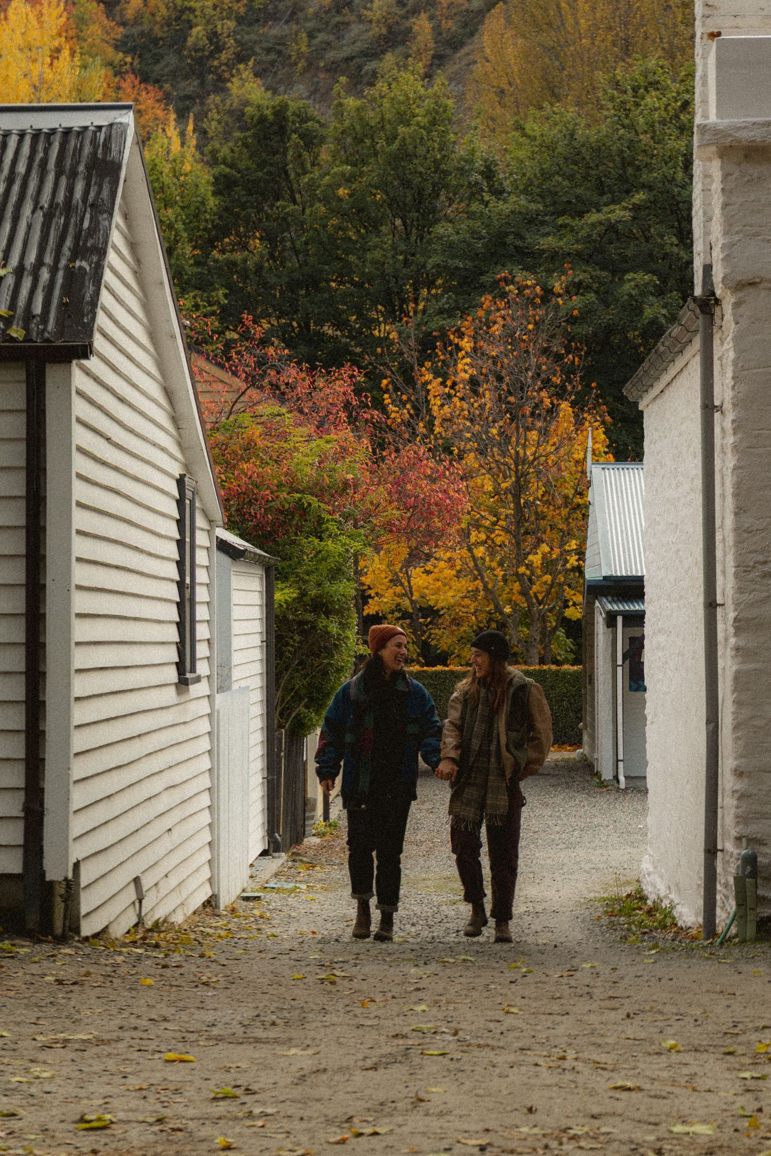 Two people strolling along a rustic cobblestone path flanked by white wooden buildings amid vibrant fall foliage.