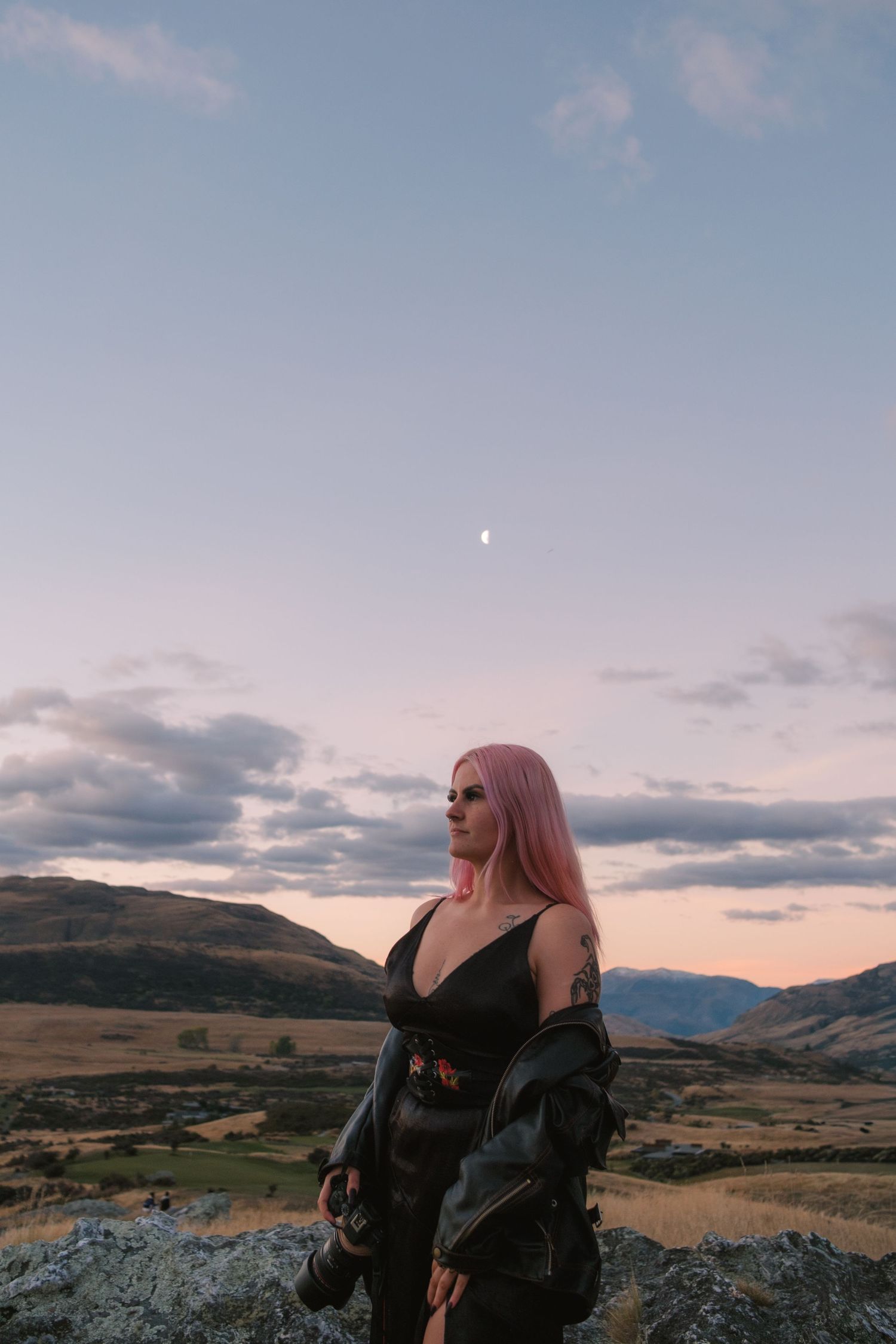 Woman with pink hair in black outfit stands on rocky hilltop at dusk with mountains and crescent moon behind her.