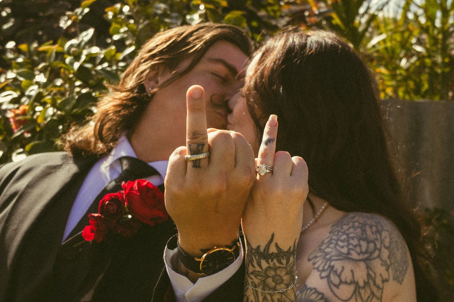 Couple kissing at wedding while flipping off camera, groom in suit with boutonniere, bride with floral tattoo sleeve.