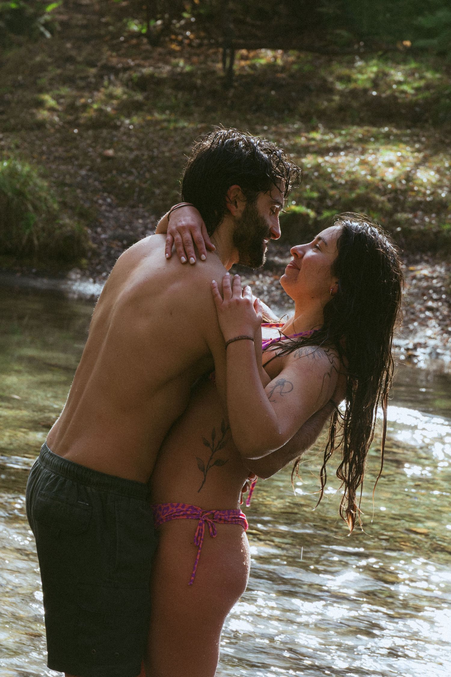 Romantic couple embracing in a shallow forest stream, wearing swimwear, with wet hair and loving gazes.