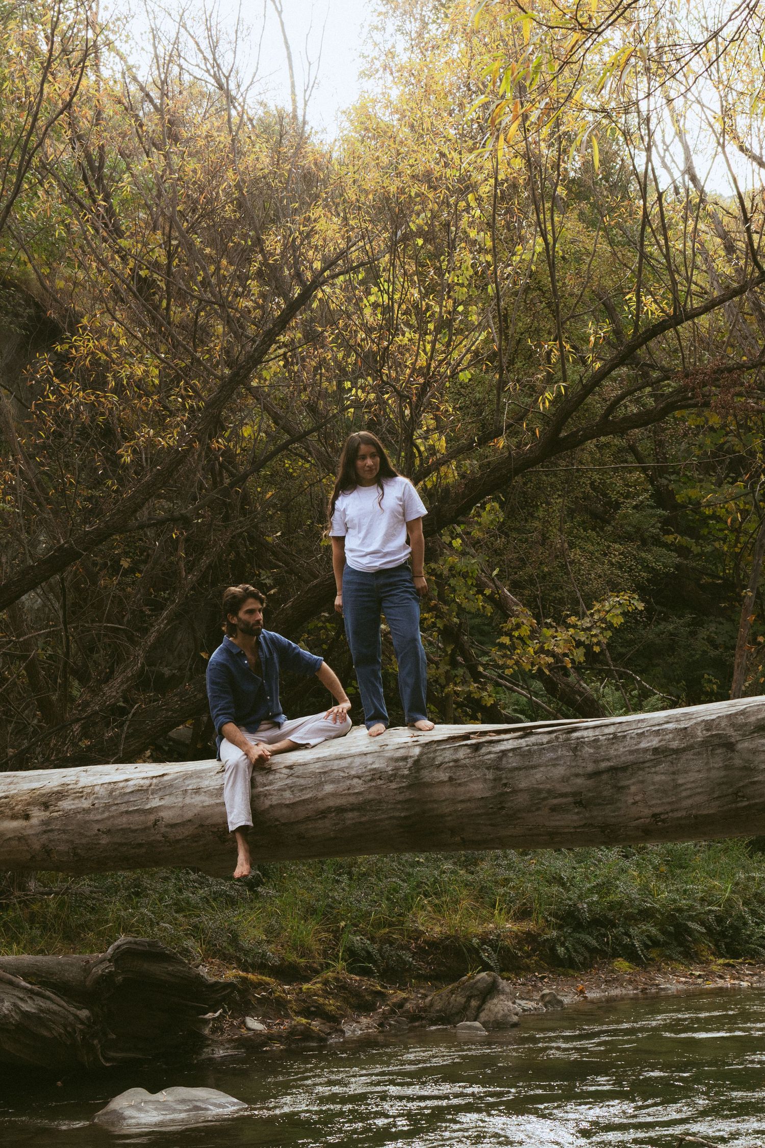 A couple relaxes on a fallen log by a river in an autumn forest, one sitting and one standing barefoot.