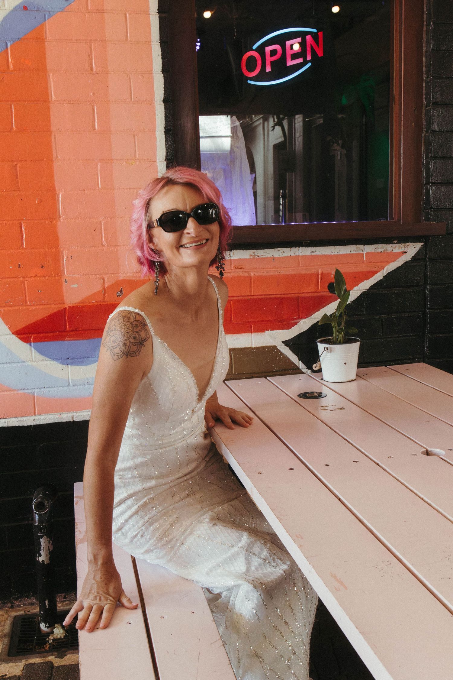 Bride wearing sunglasses sits at picnic table and smiles.