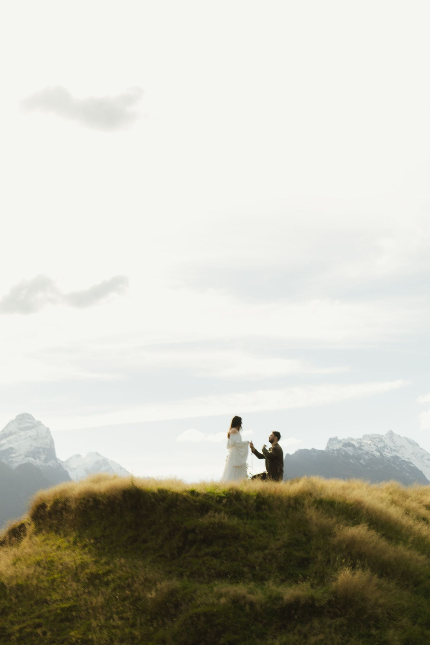A person proposes on one knee atop a grassy hill with stunning mountain peaks and cloudy sky backdrop.